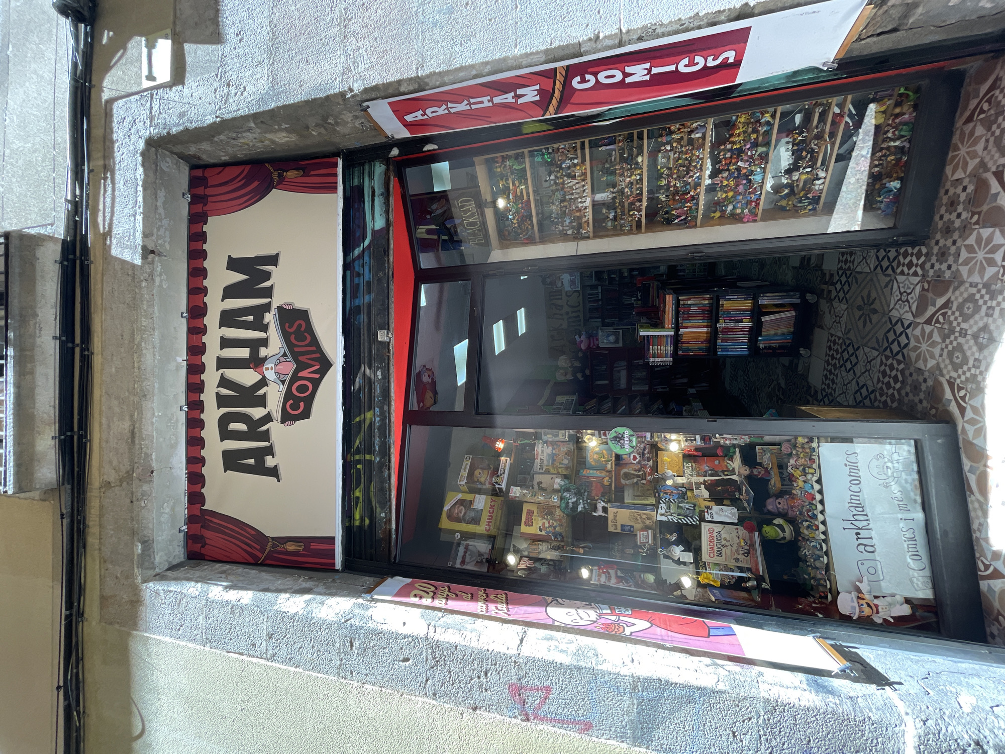 This media file captures the exterior of a comic book store named "ARKHAM COMICS" located in Barcelona, Spain. The storefront features a large, prominent sign with the store's name, rendered in bold, dark letters against a cream background, framed by red curtains. To the right of this sign, the store's entrance is visible, with large glass windows displaying an array of merchandise.
Inside the store, shelves are packed with comic books, action figures, and other collectibles. The upper portion of the display showcases numerous small figures lined up on shelves, while the lower part reveals more comic books, boxed merchandise, and promotional items. A handwritten sign on the window reads "arkhamcomics" with an Instagram logo and "Comics i més," suggesting the store offers a variety of comics and possibly other items.
The building's facade is constructed with textured concrete and stone elements, characteristic of older architecture. A decorative tiled pattern adorns the wall to the right of the entrance. The lighting suggests it's daytime, with sunlight casting shadows and highlighting the textures of the building. The overall impression is that of a vibrant, well-stocked specialty shop catering to fans of comics and collectibles.