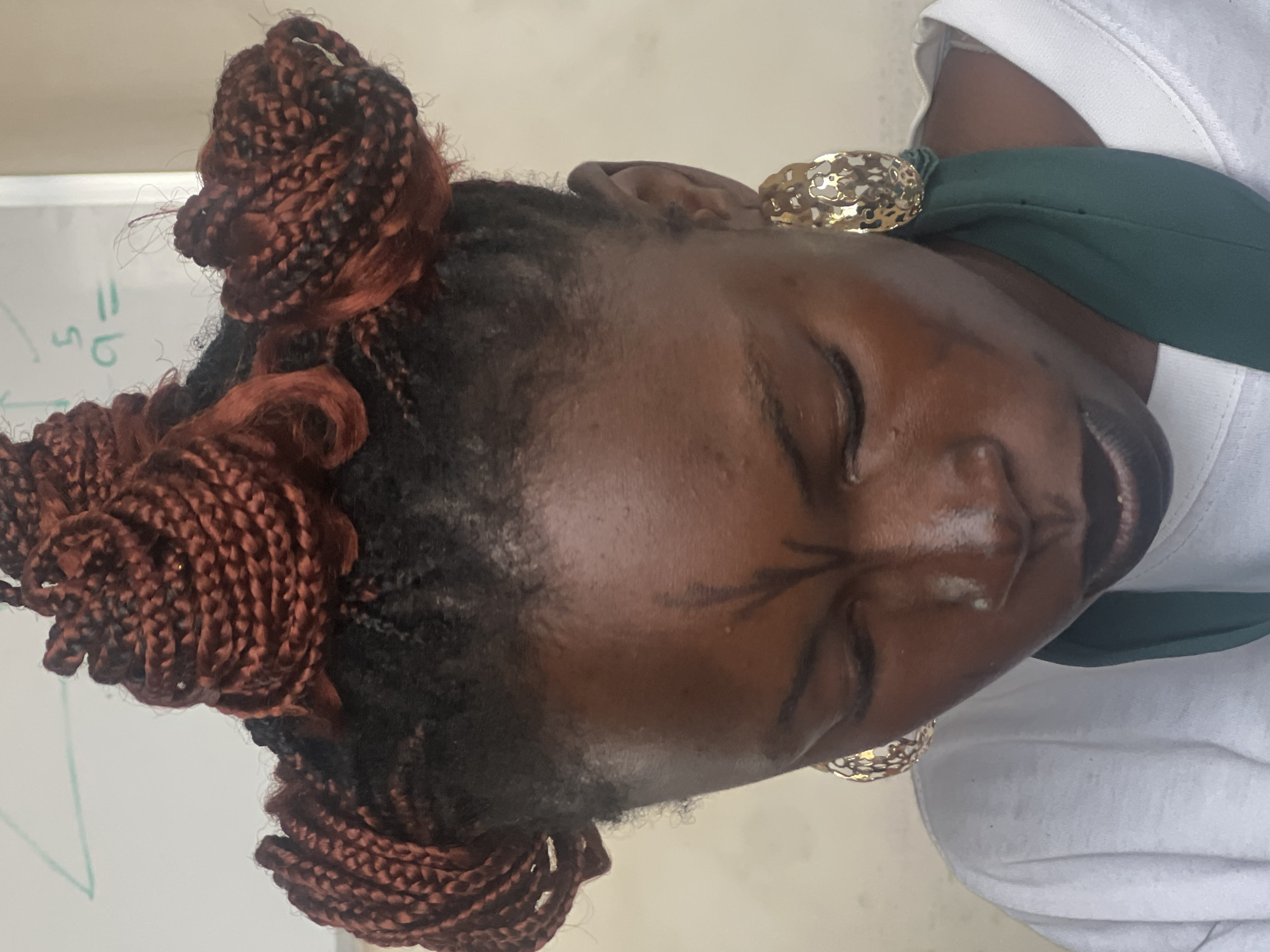 The image shows a close-up, head-and-shoulders shot of a young Black woman with dark skin, styled hair in reddish-brown braids tied into four buns, and large, gold, hoop earrings. She is wearing a white t-shirt and a dark green scarf around her neck. She has a distinctive black marking on her forehead, resembling a stylized "V" with curved lines extending downwards. The background appears to be a whiteboard with faint mathematical equations written on it. The lighting is bright, suggesting an indoor setting with natural light. The image appears to be a portrait, possibly taken in an educational or creative environment. There is no visible indication of the specific location within the city of Jos, Nigeria.