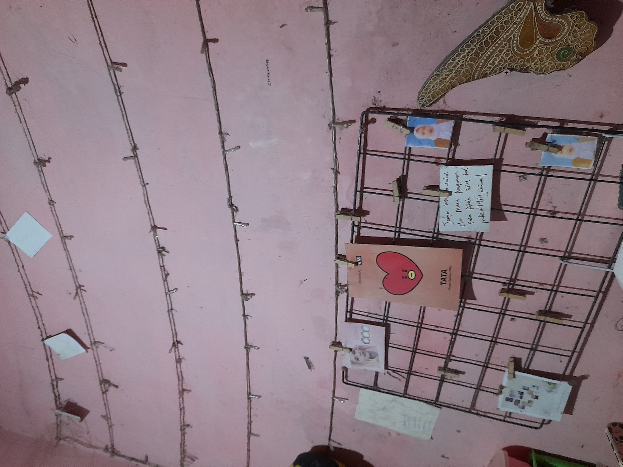 This media file depicts a section of a wall in what appears to be a bedroom or personal space, likely in Pulokulon, Indonesia. The wall is painted a soft pink and is adorned with a grid of thin wires, possibly for displaying items. Attached to these wires by small clothespins are various personal mementos.
The most prominent items on display are a series of small photographs, some showing children. There is also a card featuring a character labeled "TATA," which is a stylized red heart with eyes and a mouth, identified as "Super Curious TATA." Adjacent to this is a handwritten note, partially obscured, with text in both Latin and Arabic script, including "استغفر الله العظم" (Astaghfirullah al-azim - I seek forgiveness from Allah the Almighty) and "Jangan lupa ar-ruhul dan Misja Ampunan" (Don't forget the soul and the mission of forgiveness). Another smaller card displays a grid of tiny images.
Higher up on the wall, to the right, is a decorative item with an intricate, ornate design, possibly a piece of traditional art or handicraft, featuring earthy tones of brown and yellow.
The lighting suggests it is likely indoors, and the overall atmosphere feels personal and reflective, with a mix of photographic memories, playful character art, and religious or spiritual reminders. The presence of multiple strings of small lights, some visible running along the wires, hints that the space may be decorated with a whimsical or ambient lighting scheme. The visual texture of the wall and the various items create a sense of a lived-in, personal environment.