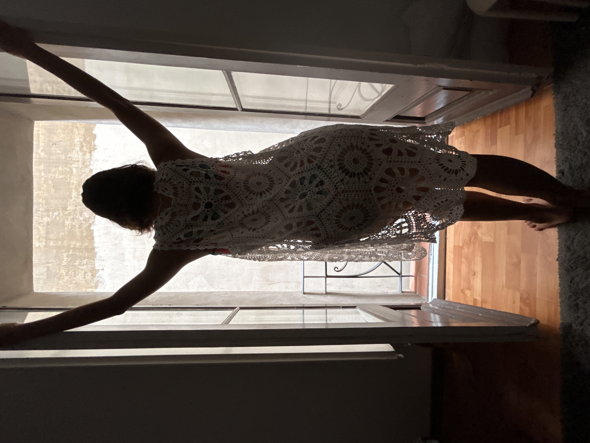 A person is visible from behind, wearing a white crocheted dress. They are standing in what appears to be an open window or doorway with a white frame. The person has their arms outstretched, touching the sides of the frame. Outside the window is a light-colored wall, possibly of a building exterior, with wrought iron details. The room inside has wooden floors and a light gray rug is visible in the corner. The image suggests an indoor-outdoor setting, though the exact location in Barcelona cannot be determined from the visual information provided.