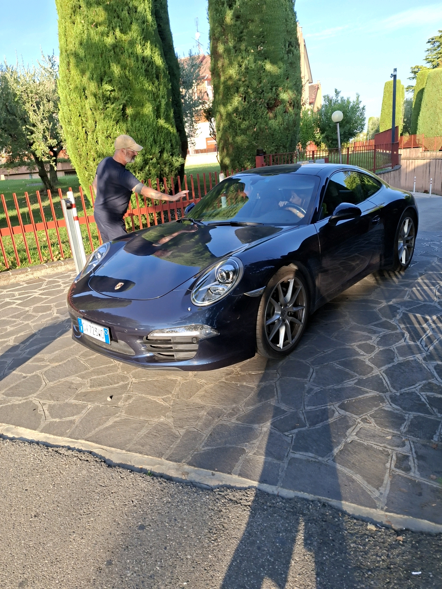 In the city of Sirmione, Italy, a dark blue Porsche 911 Carrera S is parked on a paved driveway with a hexagonal stone pattern. The car's license plate reads "CV 723WD". A man wearing a dark t-shirt and a beige baseball cap is standing next to the driver's side door, with his hand resting on the car. He appears to be interacting with the vehicle or its occupant. Inside the car, a person can be seen in the driver's seat, with their hands on the steering wheel. The background features tall green cypress trees, a red metal fence, and a building with terracotta roof tiles, suggesting a residential or private property setting. The lighting indicates it is daytime, with sunlight casting shadows on the ground.