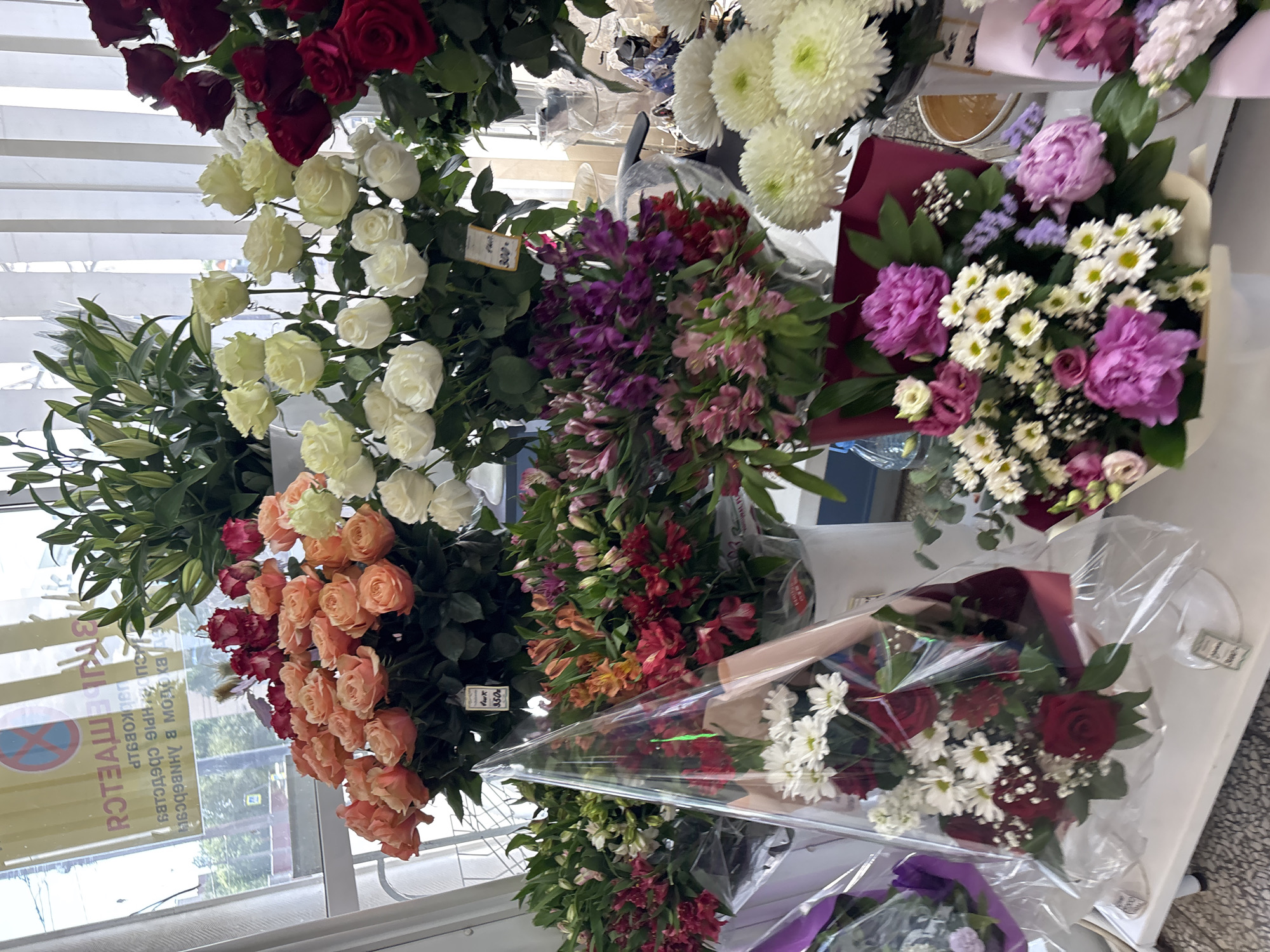 The image captures a vibrant display of various flowers in what appears to be a flower shop or stall, seen from a slightly elevated perspective. The scene is dominated by numerous bouquets and bunches of fresh cut flowers in a wide array of colors.
The primary subjects are the flowers themselves, which include large quantities of roses in deep red, white, and peach/orange hues. Other prominent flowers are white chrysanthemums with green centers, purple and pink alstroemeria, purple peonies, delicate white baby's breath, and green lily leaves with buds. The flowers are arranged in large bunches, some standing upright, while others are prepared as bouquets wrapped in clear plastic or colored paper, such as maroon and purple.
The activity depicted is the presentation and sale of flowers. Many of the flower bunches have small price tags attached; one clearly shows "350р" (350 rubles).
Notable details include the bright, natural daylight illuminating the scene, suggesting it is daytime. A large window or glass display on the left reflects the exterior, showing parts of a street or outdoor area. The reflections also reveal Cyrillic text from various signs. Partially visible words include "ЗАПРЕЩЕНО" (Forbidden/Prohibited), accompanying a no parking/stopping sign (red circle with diagonal line). Other reflected text includes parts of words like "В АННАБЛЕССИ" (In Annablessi), "ЦВЕТЫ" (Flowers), and "УНИВЕРМАГ" (Department store), consistent with the provided location context of Russia. The overall impression is one of abundance and freshness.