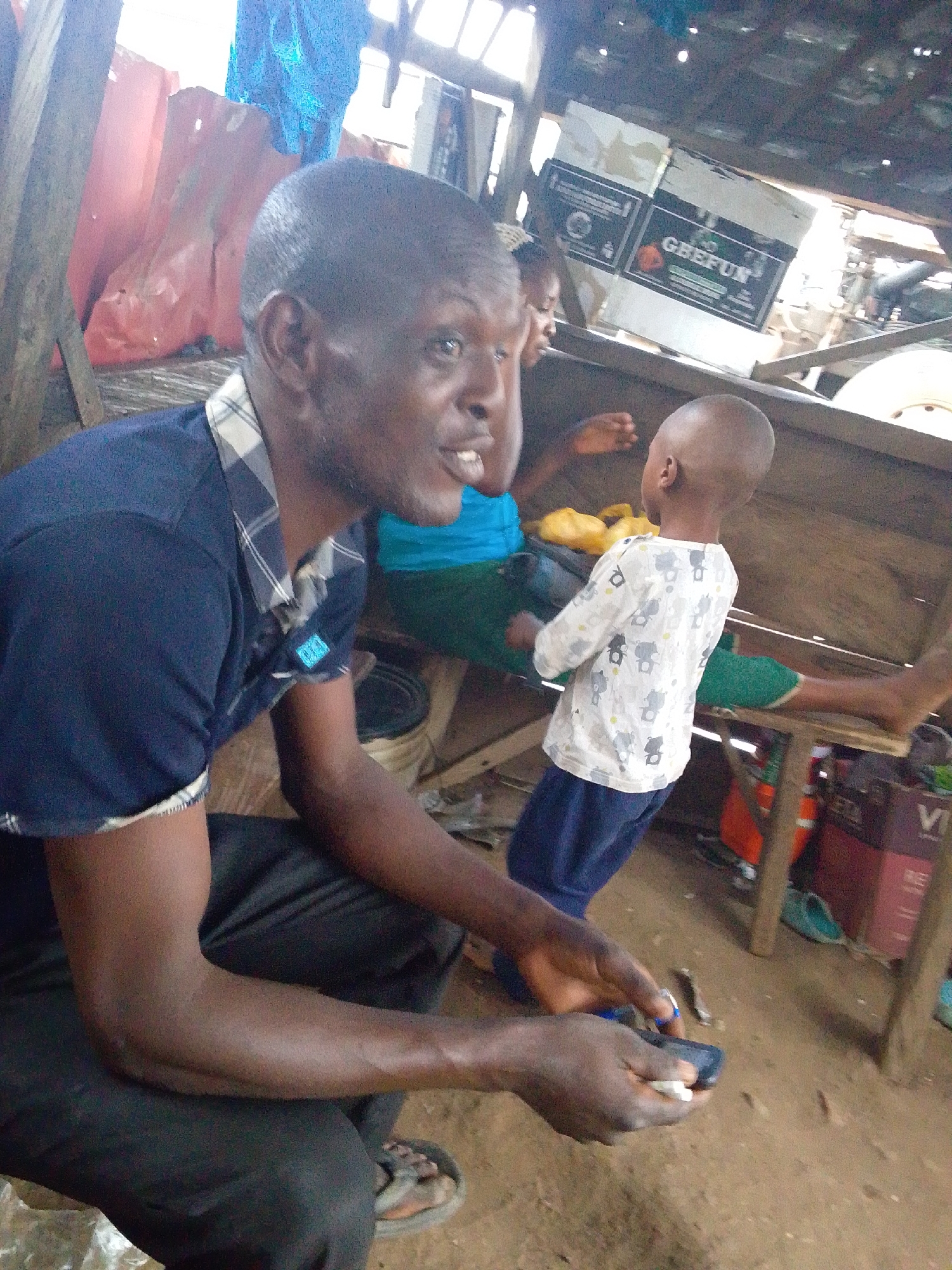 This image captures a vibrant, informal indoor scene in Lokoja, Nigeria, illuminated by natural daylight.
In the foreground, a dark-skinned man with short hair is squatting or sitting low. He wears a dark blue polo shirt with a plaid collar and rolled-up sleeves, and dark trousers or shorts, with sandals on his feet. His mouth is open, suggesting he is speaking animatedly or reacting with surprise, and his expression is engaged. He holds a small, dark object, possibly a mobile phone, and a small white object in his hands.
To the man's right, a young child with a bald head stands facing away from the camera. The child wears a light-colored long-sleeved shirt patterned with grey elephants and dark blue pants. The child appears to be observing the man or something to the left.
Further in the background, another person, possibly a child, is partially visible. They are seated on a wooden bench or platform, wearing a bright blue top and green bottoms. The setting appears to be a rustic structure, perhaps a market stall or a communal area, built with wooden beams and planks. The ground is a light brown dirt surface.
Various objects are scattered in the background, including red and blue tarps or fabrics, and cardboard boxes. One prominent white box in the upper right background clearly displays the text "GEEFUN." Another box, red, is partially visible near the bottom right with "VITA" printed on it. The overall impression is one of everyday life in a simple, community-oriented environment.