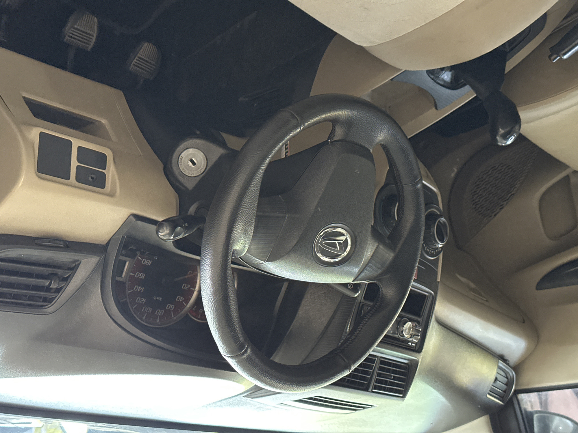 This is a close-up, eye-level interior shot of a car's dashboard and steering wheel, taken in Bulakrejo, Indonesia. The car's interior is primarily beige and black. A black steering wheel with the Daihatsu logo is prominently featured in the center. To the right of the steering wheel is a speedometer and tachometer cluster, with the speedometer showing speeds up to 180 km/h. A black gear shift lever is visible to the left of the steering wheel. The dashboard has air vents and several buttons and switches. The seats appear to be upholstered in beige fabric. The overall impression is of a well-maintained, standard passenger vehicle interior.