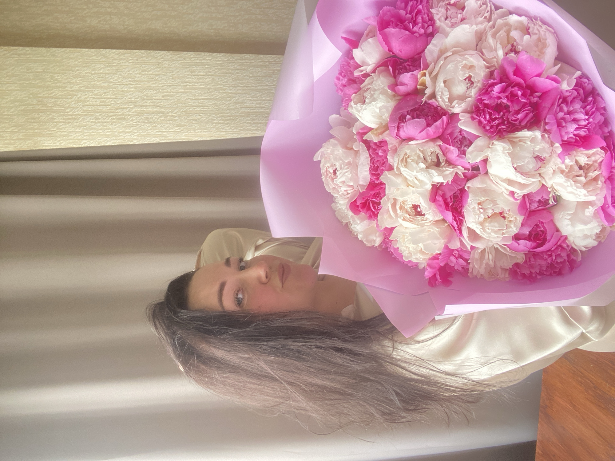 A woman with long dark hair is holding a large bouquet of pink and white flowers wrapped in pink paper. She is wearing a light-colored, silky shirt. The background includes a textured wall and a wooden surface. The image is indoors and appears to be taken in a residential setting. The city of Sochi, Russia, is not discernible from the visual elements of the image.