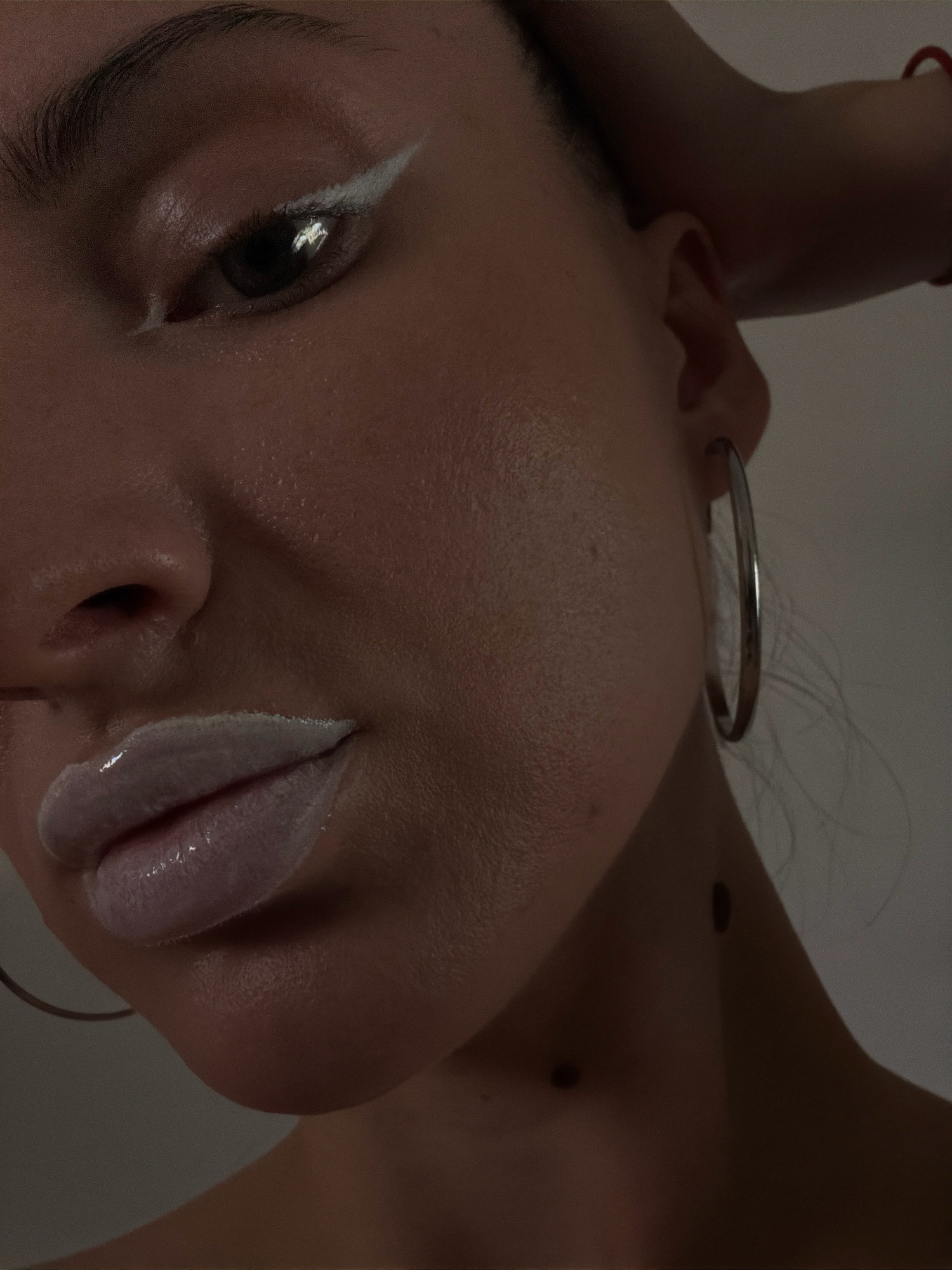This is a close-up, low-angle, medium shot of a person's face, with a focus on the eye and lips. The individual has fair skin and is wearing dramatic white eyeliner and light-colored, glossy lipstick. They are also wearing a large hoop earring. A portion of their neck and shoulder is visible, with two small dark marks on the skin. The lighting is dim, creating shadows and highlighting the contours of the face. The background is a plain, light-colored surface. The image is a portrait, likely taken indoors, with no contextual clues indicating a specific location in Sharm El Sheikh, Egypt, or any other city.