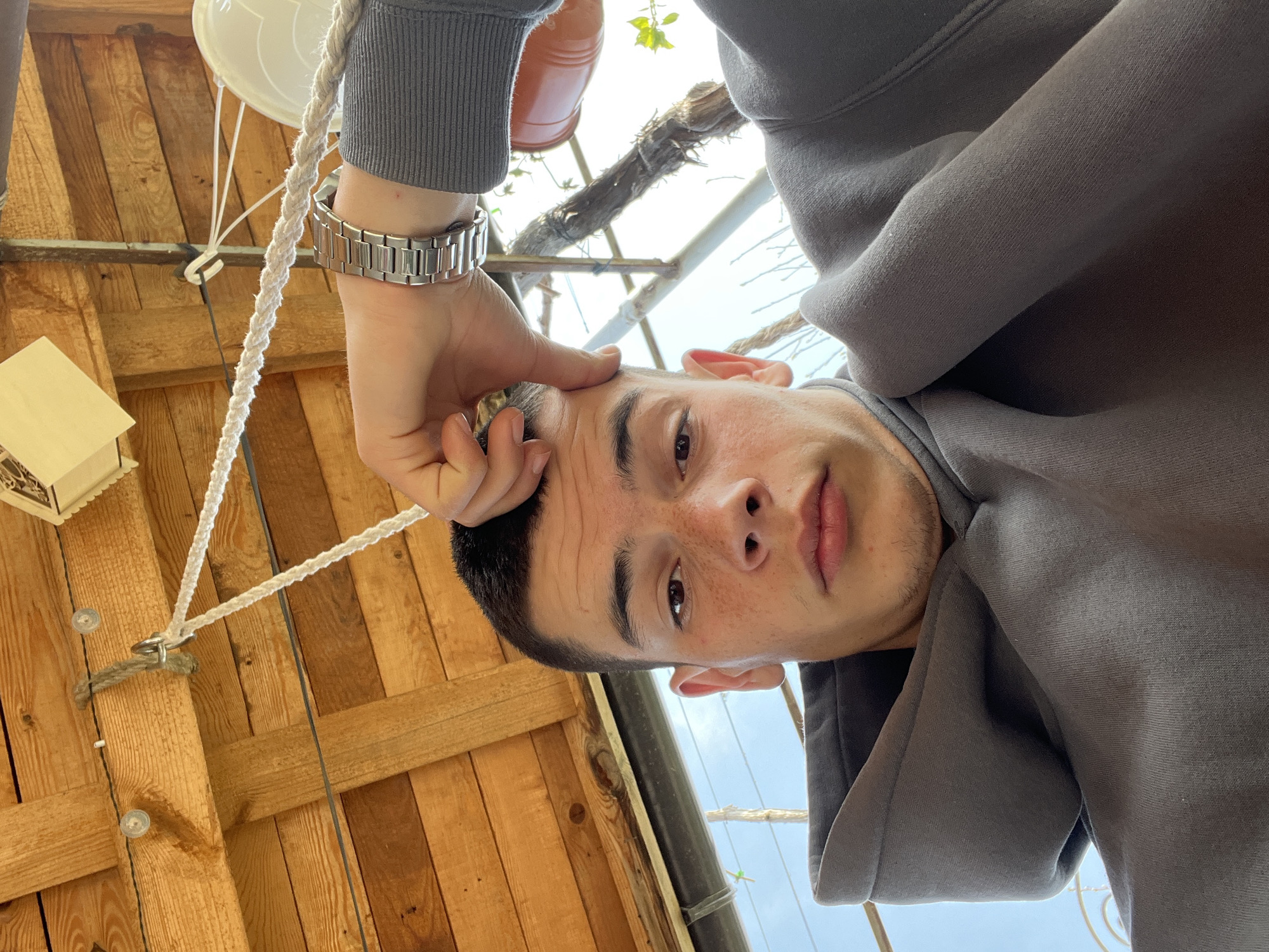A tilted selfie shot from a low angle of a young man in a grey hooded sweatshirt. He is touching his forehead with his left hand, which has a silver watch on the wrist. The background shows a wooden structure with planks and rope, suggestive of a rustic or unfinished interior, along with glimpses of greenery and sky visible through the structure. This could be an outdoor structure or an unfinished building in a rural setting. The location is stated as Karavelovo, Bulgaria.