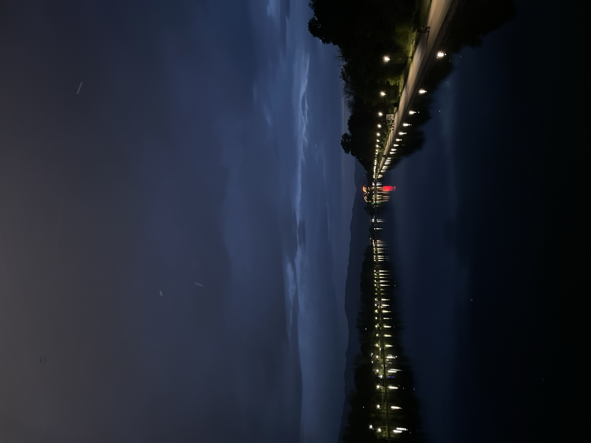 The image shows a night scene in Plovdiv, Bulgaria, specifically depicting the Rowing Canal. A row of lights illuminates a path along the canal, with their reflections visible in the dark water. In the distance, there are illuminated structures with a distinct red glow, possibly buildings or decorative features near the canal. The sky is dark, suggesting a late evening or night, with visible clouds. The trees on the side of the path are silhouetted against the sky. There's minimal human activity immediately visible.