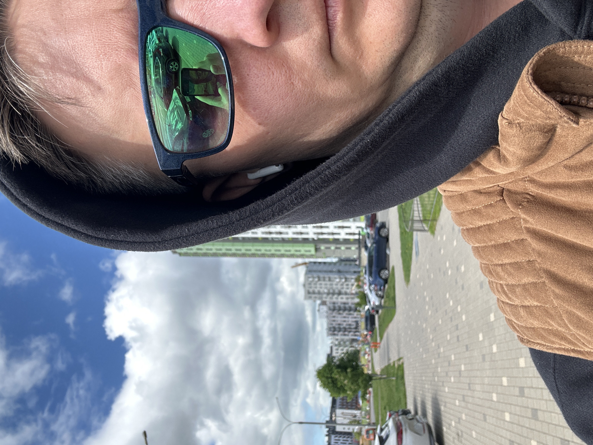 A distorted first-person perspective photo shows a person wearing sunglasses with green lenses, reflecting the surroundings. The individual is also wearing a black hoodie with a brown suede jacket. The background displays a cityscape under a cloudy sky. Modern high-rise buildings, parked cars, trees, and a paved sidewalk are visible. The location is identified as Бараўлянскі сельскі Савет, Belarus. The person is holding a phone, also visible in the sunglasses' reflection.
