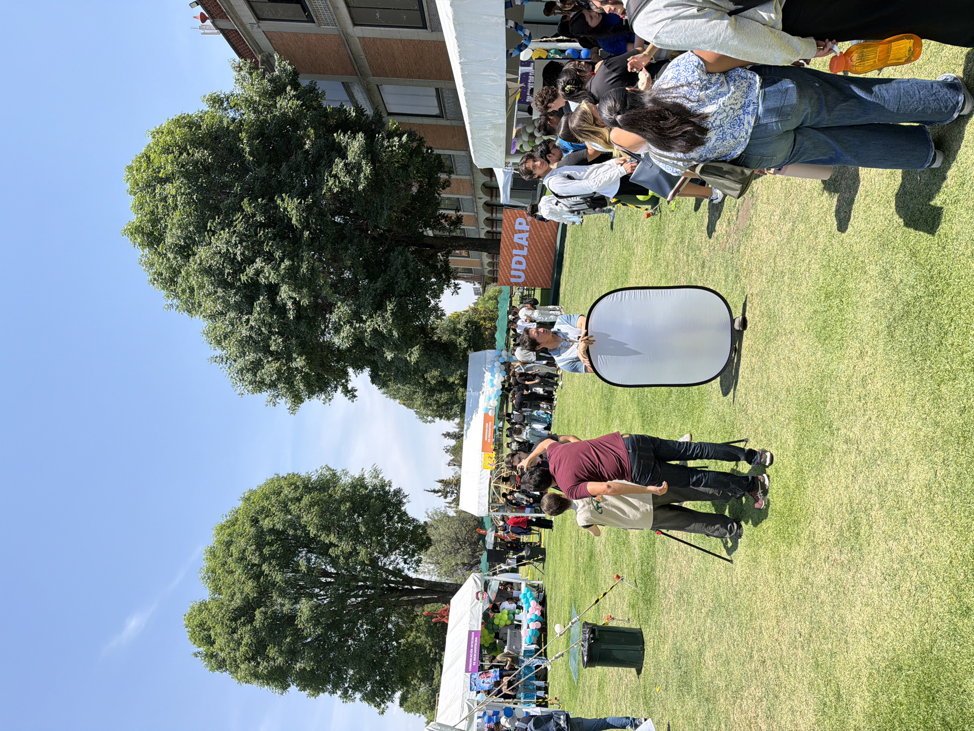 This media file captures an outdoor event on a bright, sunny day at what appears to be a university campus in San Andrés Cholula, Mexico. The image is rotated counter-clockwise by 90 degrees.
Numerous people are gathered on a large, well-maintained grassy area. In the foreground, two individuals are seen with a large, white, circular photography reflector, suggesting a photoshoot or media-related activity. One person holds the reflector upright, while another, wearing a maroon shirt and dark pants, is bent over, possibly setting up equipment or adjusting something.
Several large, white canopy tents are set up across the green lawn, indicating an outdoor fair or exhibition. People are congregated around these tents, some standing and interacting, others seemingly browsing displays or engaging with booths. Balloons in shades of light blue, white, and pink decorate some of the tents, adding to a festive atmosphere.
Visible text on banners identifies key aspects of the event. A prominent orange banner in the mid-ground clearly displays "UDLAP," referring to the Universidad de las Américas Puebla, which confirms the location context of San Andrés Cholula. Another partially visible banner on a white tent reads "COMUNICACIÓN ESTRATÉGICA Y PRODUCCIÓN DE MEDIOS," likely indicating a university department or program showcasing its work. A third banner also mentions "INNOVACIÓN Y PRODUCCIÓN DE..." (likely 'PRODUCTOS').
In the background, multi-story buildings with brick facades are visible, along with large, leafy green trees under a clear blue sky, reinforcing the campus setting. The bright sunlight suggests the event is taking place during midday or early afternoon.