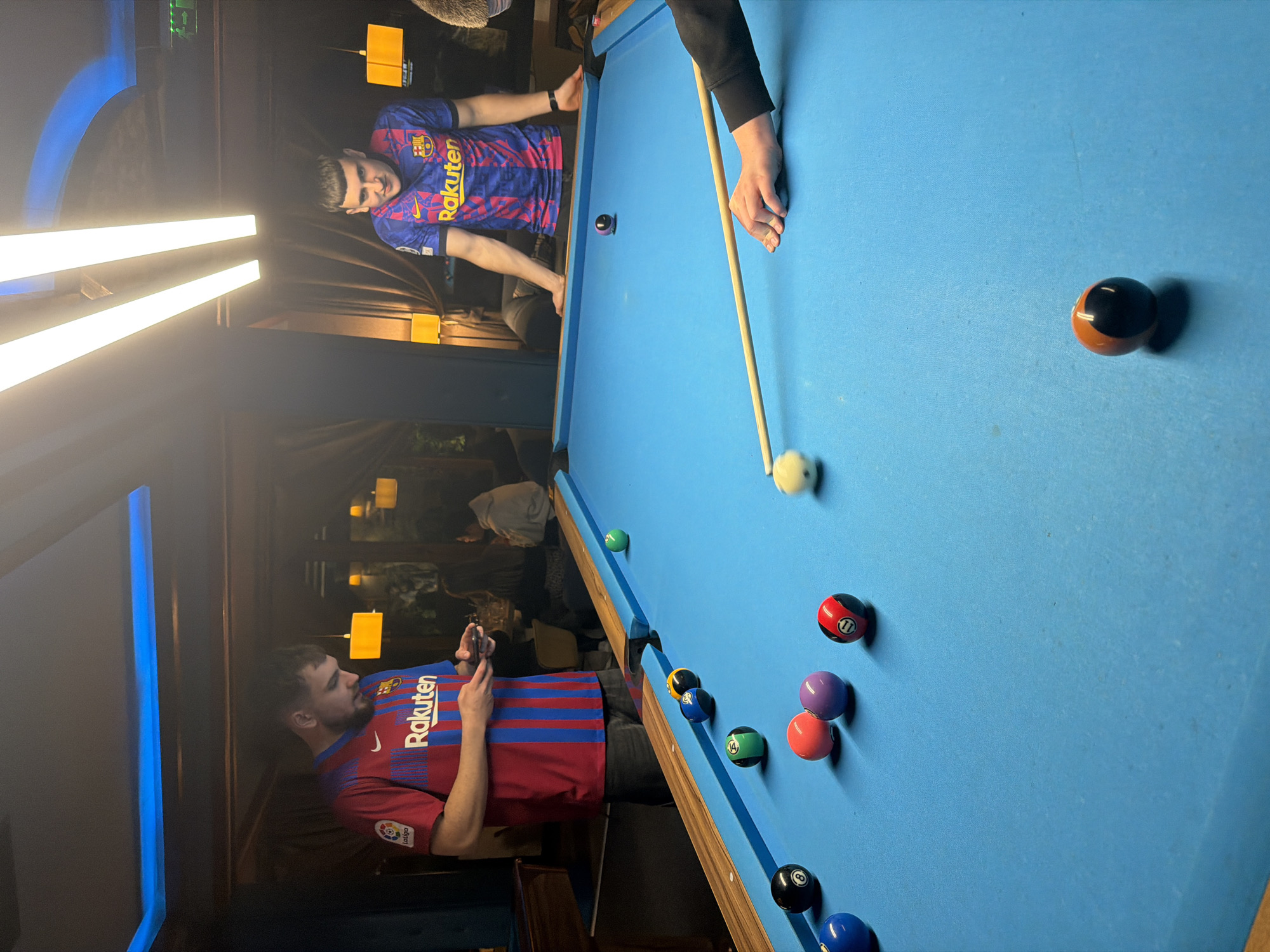 The image depicts a billiards game in progress. Two individuals wearing Barcelona football club jerseys are present. One is holding a smartphone, while the other is poised to strike the cue ball. A cue stick is visible. Several billiard balls are scattered on the blue-felted pool table, including the cue ball, a black 8-ball, and various other numbered balls. The scene appears to be indoors, possibly in a bar or pub setting. The location is Plovdiv, Bulgaria. The lighting is a mix of overhead fluorescent and ambient sources.