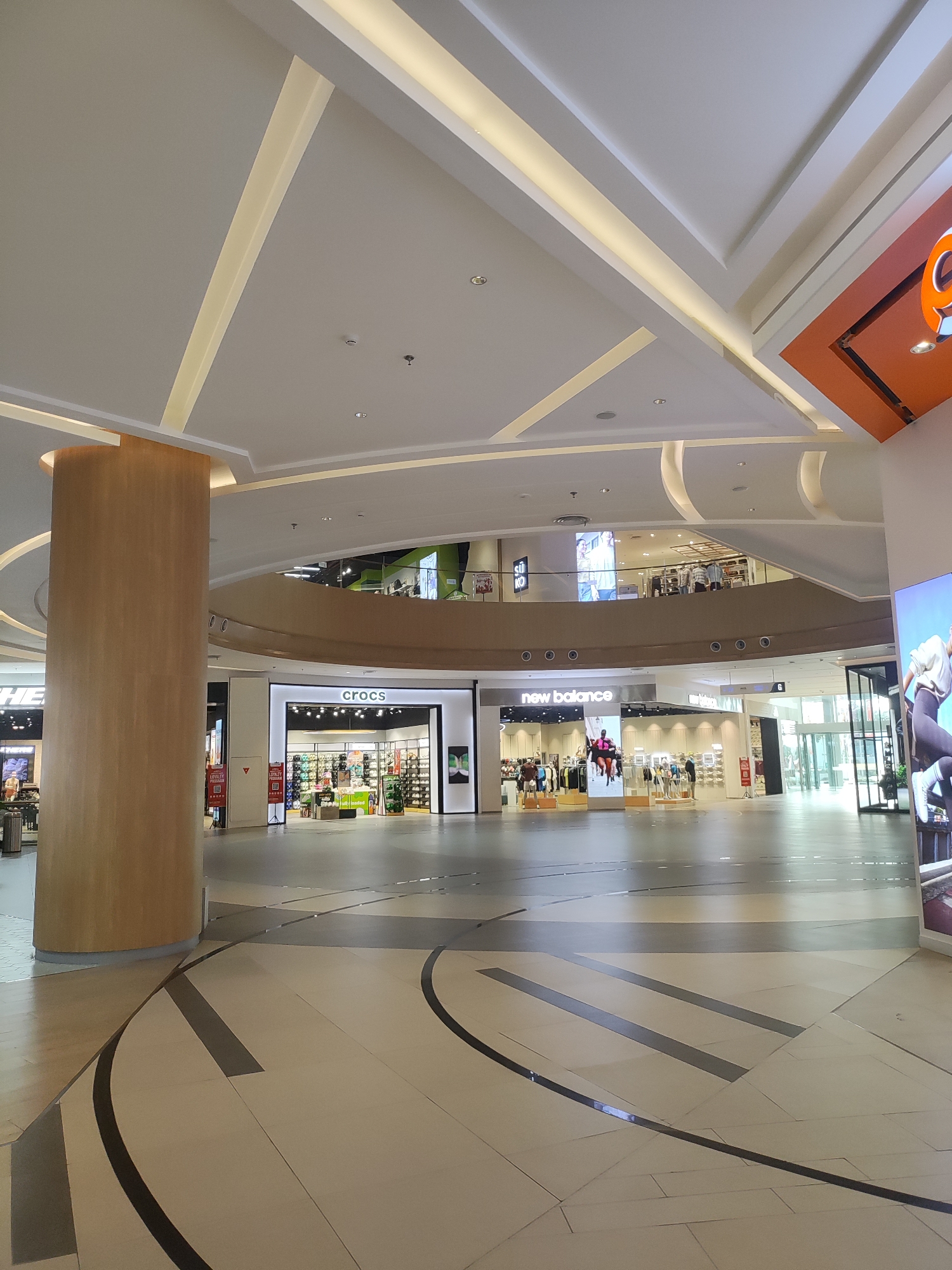 This is an indoor shot of a modern shopping mall. The image captures a wide view of the mall's interior, featuring multiple levels and storefronts. In the foreground, a large, cylindrical wooden pillar stands to the left. The floor is tiled in a neutral color with dark, geometric patterns.
Several shops are visible. To the right of the pillar, a "Crocs" store is clearly marked with its logo and displays rows of footwear. Further to the right, a "New Balance" store is visible with mannequins showcasing athletic wear and displays of shoes. Above these ground-level stores, a second level is visible with more shops and people browsing. In the upper right corner, a large orange sign with a partial "Q" logo is present. The mall's ceiling is complex, with white architectural features and integrated lighting, creating a contemporary and spacious atmosphere. The lighting appears to be a mix of ambient and spotlighting, illuminating the shops and walkways. The overall scene suggests a well-maintained and active commercial space, likely located in a city like Lambangjaya, Indonesia, given the context. There are no prominent individuals or specific actions depicted.
