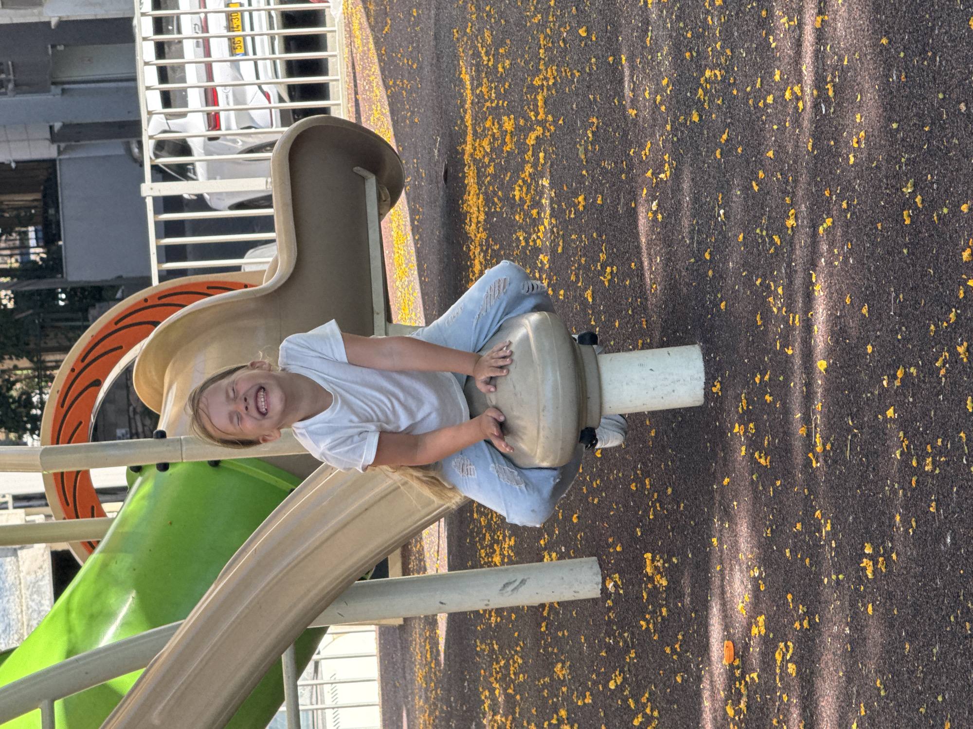 A young girl with blonde hair is sitting on a playground structure, smiling with her eyes closed. She is wearing a white t-shirt and light blue jeans with rips on the knees. The playground equipment is beige and green, and there are yellow flower petals scattered on the dark brown ground. In the background, a white car is visible behind a white fence. The scene appears to be an outdoor public space in a city.