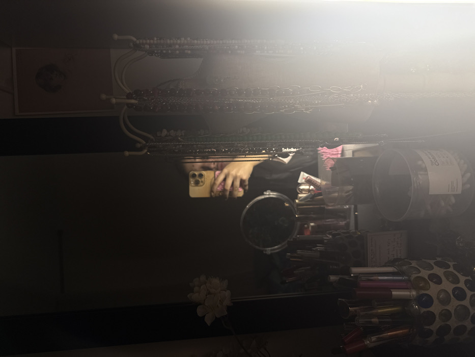 A mirror reflects a cluttered space: a person holding a phone, makeup containers, tubes, and a small round mirror. Above the mirror, jewelry hangs from a rack. A framed photo is visible on the left. In the foreground, a flower arrangement sits, partially obscured. The scene is dimly lit, with a bright light source at the top.