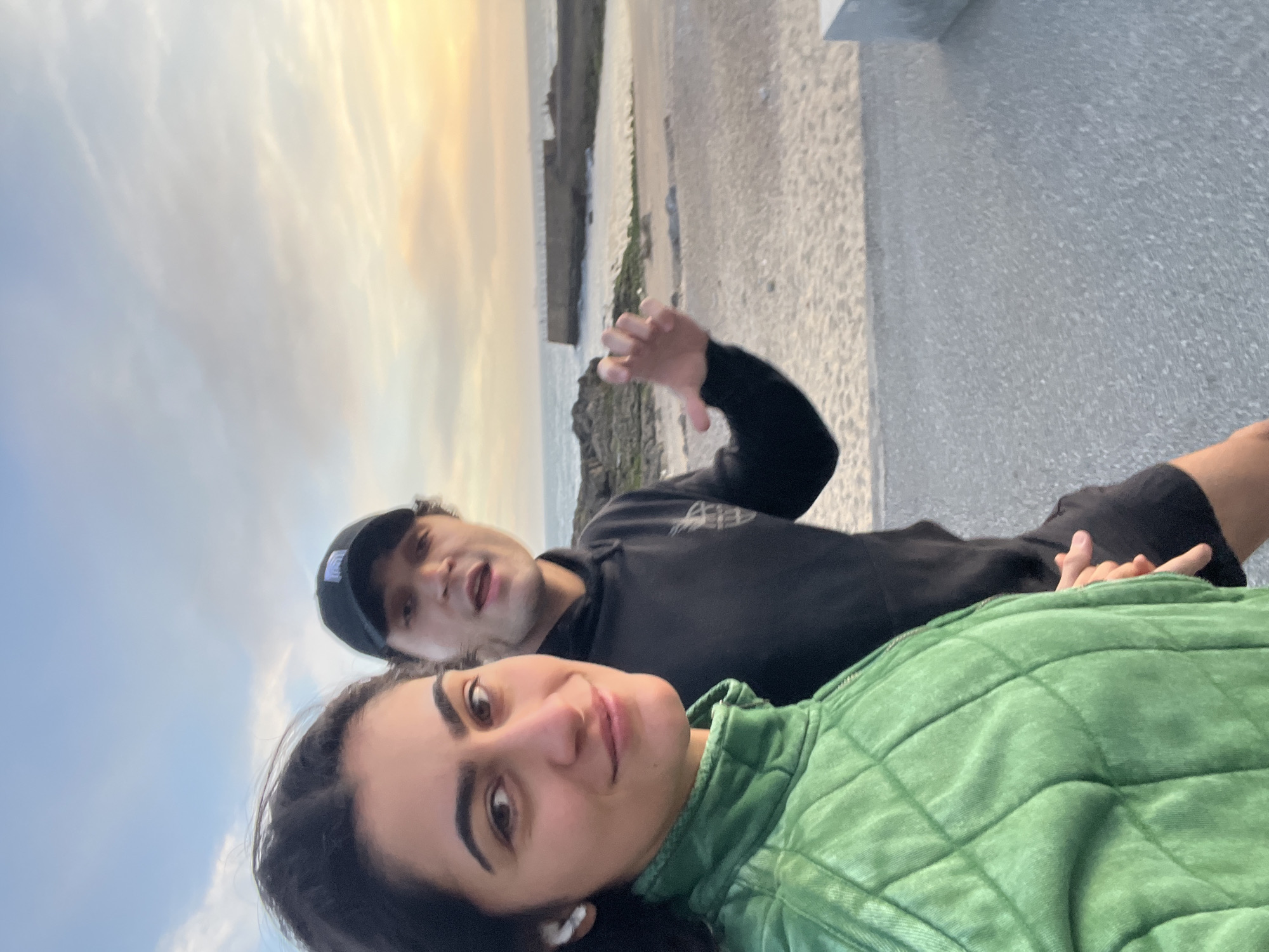 Two individuals are posing for a selfie. The background shows the shoreline, ocean, and a cloudy sky. The location is likely Porto, Portugal, given the architecture and coastal setting. A woman in the foreground is wearing a green quilted jacket and has dark hair. Behind her is a man wearing a black hoodie and a baseball cap. The man is gesturing with his hand. They are standing on a concrete surface.