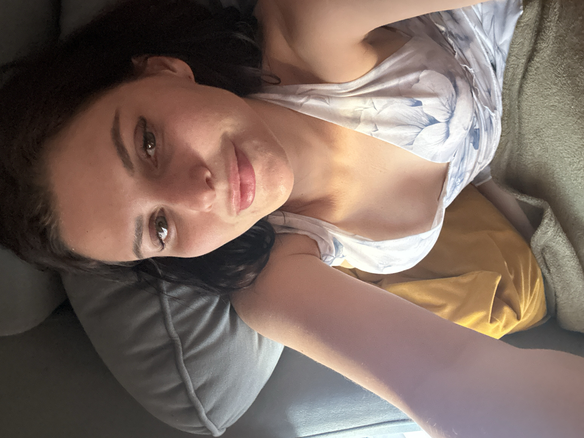 A young woman with dark hair and brown eyes is lying down on a sofa, looking at the camera. She is wearing a white dress with a blue floral print. The lighting in the image suggests it was taken indoors, possibly in a living room. The woman's arm is extended towards the camera, indicating she might be taking a selfie. The background shows the sofa cushions and a folded blanket.