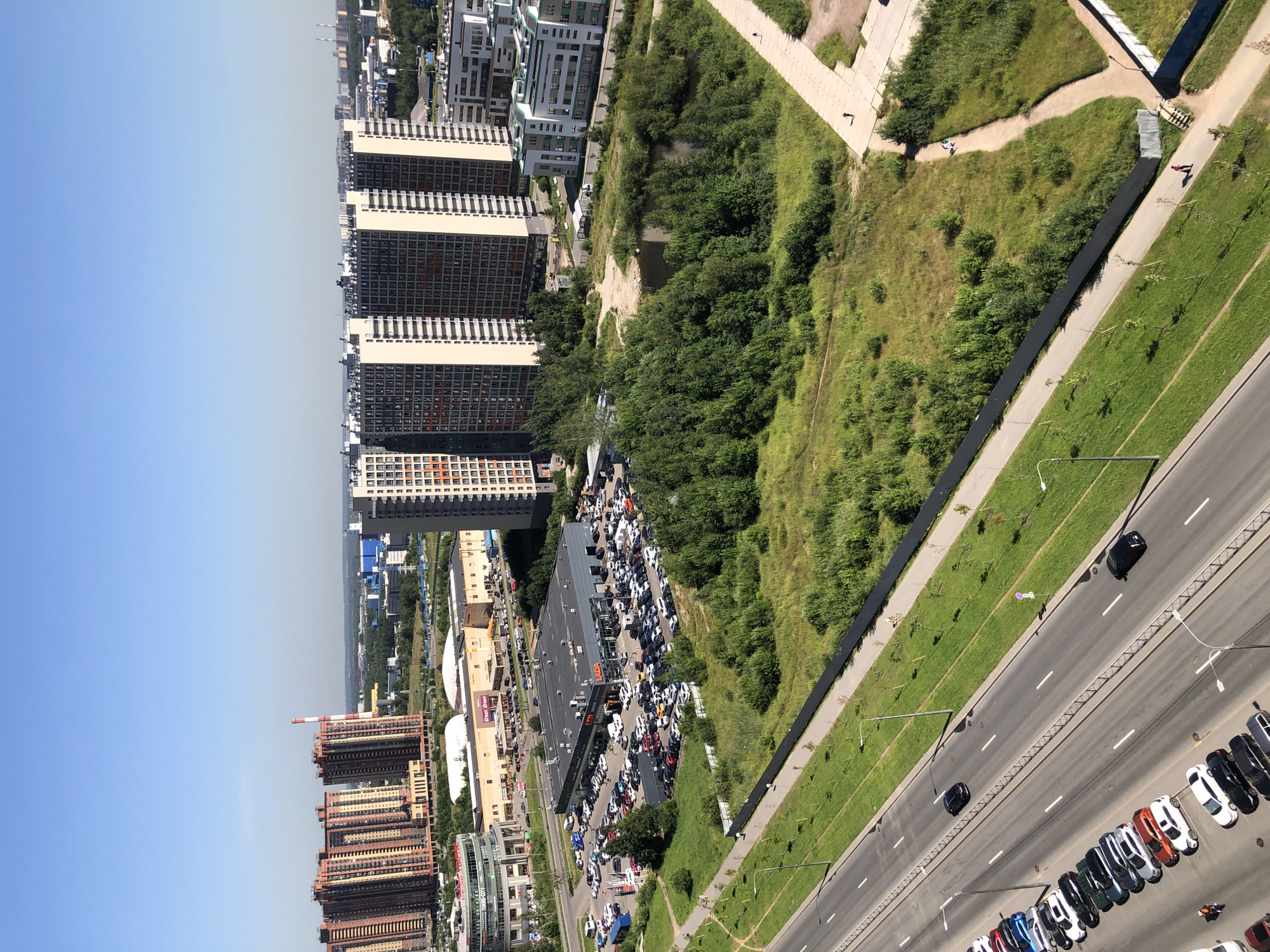 This is an aerial, landscape-oriented photograph taken from a high vantage point, likely a tall building, looking down and across a suburban or urban landscape under a clear, bright sky, suggesting daytime. The scene is dominated by a mix of modern residential buildings and green spaces, with a busy road featuring parked and moving vehicles.
In the foreground, a multi-lane road runs diagonally through the image, with numerous cars parked along its sides and a few in motion. The cars are varied in color and type, indicating a populated area with significant vehicle traffic. To the left of the road, a long, dark strip, possibly a service road or a protected area, runs parallel. Beyond that, a grassy area with scattered trees and shrubs transitions into a densely vegetated hillside.
Further into the scene, a large, modern building, possibly a commercial establishment or a car dealership, is visible with many cars parked in front of it. This building has a prominent "LADA" sign displayed on its facade, identifying it as a car dealership. Beside it, a large open field or park area stretches out, bordered by more residential buildings.
The background is filled with a cluster of tall, multi-story apartment buildings, characterized by their repeating patterns of windows and balconies. These buildings are a mix of light-colored facades with darker window frames, and some exhibit reddish-brown exteriors. The architecture suggests a relatively new development.
There are a few people visible in the image, most notably two individuals walking on a paved path in the green area to the right of the commercial building, and another person with a red shirt walking on a dark path near the edge of the frame on the right. The overall impression is one of a sunny day in a developed area that combines urban living with recreational green spaces and significant car culture.