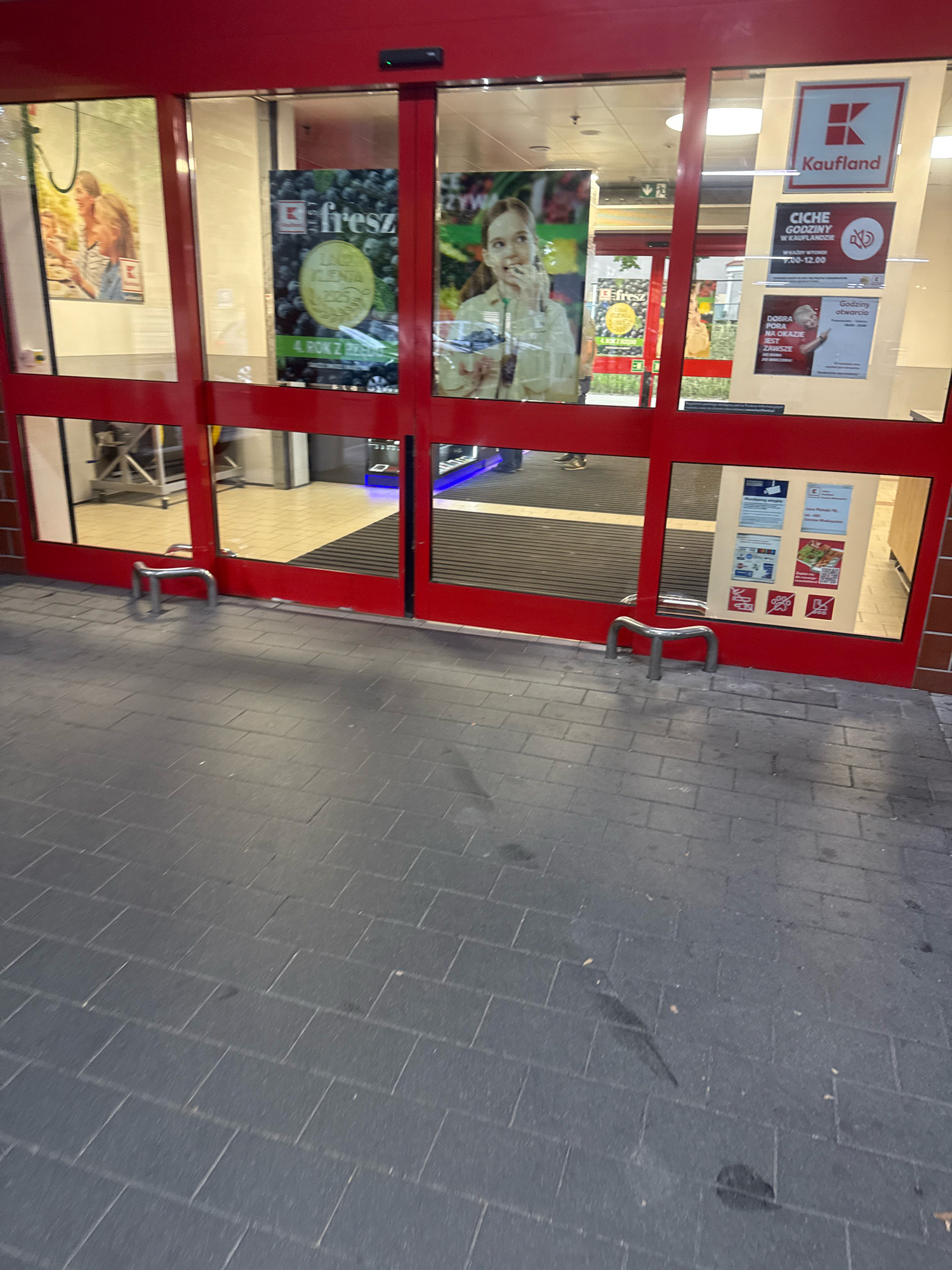 The image shows the exterior entrance of a Kaufland supermarket in Gorzow, Poland. The entrance features automatic sliding glass doors with red framing. Above the doors, signage displays the Kaufland logo and name. On the doors themselves, various posters are displayed, including advertisements for fresh produce and store hours. One poster, "CIche Godziny w Kauflandzie," indicates "Quiet Hours in Kaufland," with specific times listed from 7:00 AM to 12:00 PM. Another poster, "Godziny otwarcia," displays the opening hours.
Inside the store, visible through the glass, are shopping carts and aisles. The floor in front of the entrance is paved with dark gray rectangular tiles. Two metal U-shaped bollards are positioned on the pavement just outside the doors. The lighting suggests it is daytime, though possibly overcast, as there are reflections on the glass doors. The overall scene depicts a typical retail entrance environment.