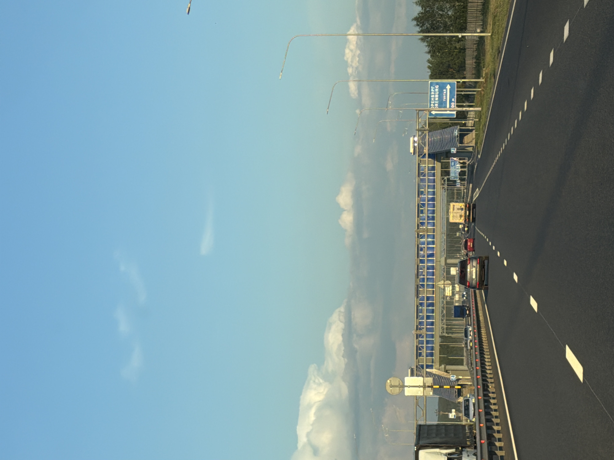 The image shows a highway in Zimnyaya Gorka, Russia, under a partly cloudy blue sky. The perspective is from the driver's seat of a vehicle moving along the highway. In the foreground, the asphalt road has white dashed lane markings. On the left side of the road, a metal barrier separates the lanes from the shoulder, where a large white truck is visible. Further down the highway, other vehicles, including cars and trucks, are traveling in the same direction.
Overhead, a pedestrian bridge crosses the highway. Blue panels are visible on the bridge's structure. Several lampposts line the right side of the highway, with trees and a fence visible beyond them. A blue road sign with white text and an arrow points to the right, indicating directions to Orenburg and Turgay. The overall scene depicts a typical highway environment in Russia during daylight.