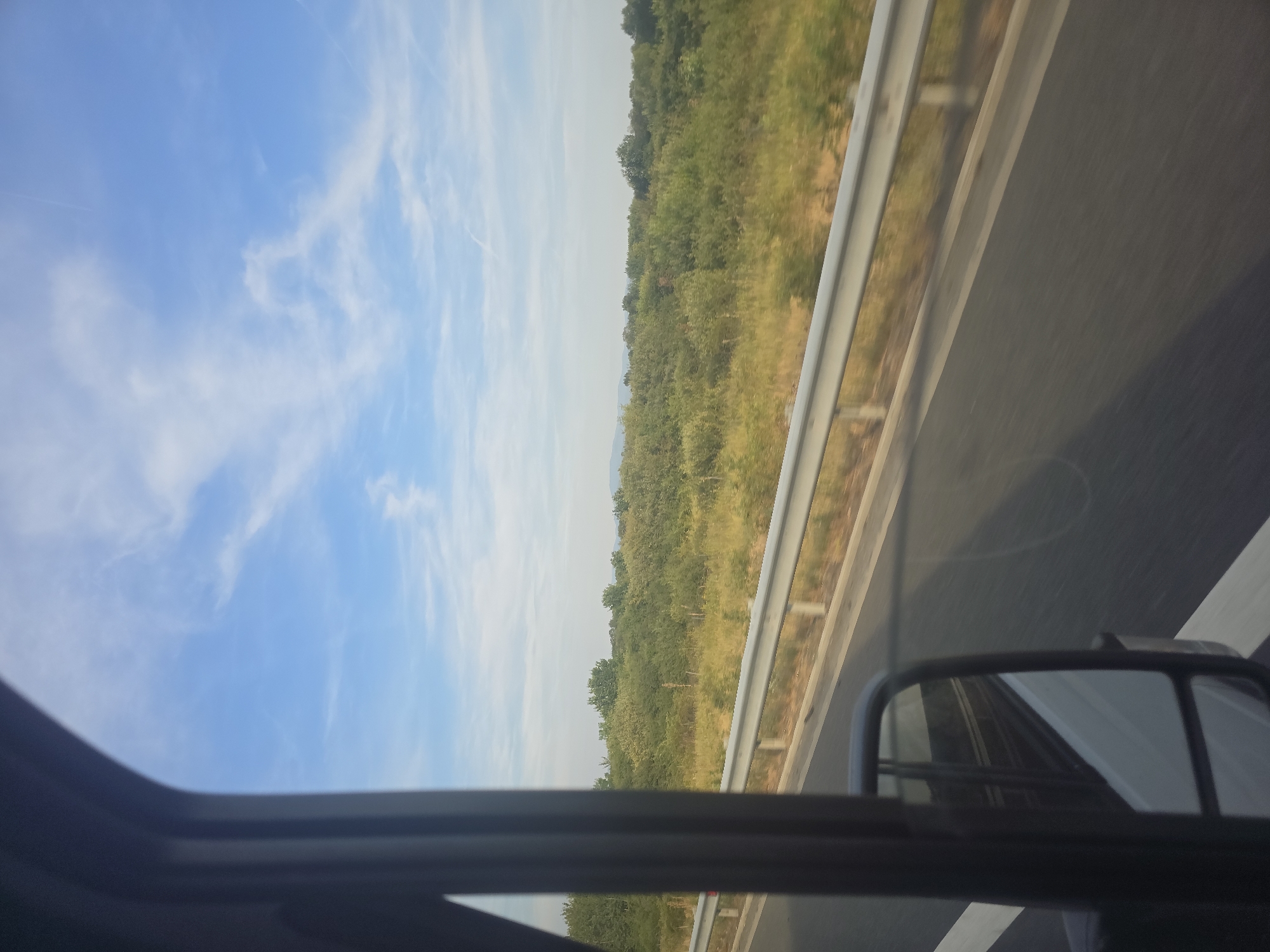 The image captures a view from a vehicle moving along a road, likely a highway, with a scenic landscape on the right. The foreground is dominated by the interior of the vehicle, specifically the dark frame of the window and a side mirror reflecting a white vehicle. The road surface is asphalt with white lane markings. To the right of the road, a metallic guardrail separates it from a field of dry grass and then a dense expanse of green vegetation, including trees and bushes. In the background, the sky is a bright blue with wispy white clouds. The overall scene suggests a journey through a rural or semi-rural area, possibly in the vicinity of Cutina, Romania, given the geographical context. There are no people visible, and the focus is on the natural environment and the road.