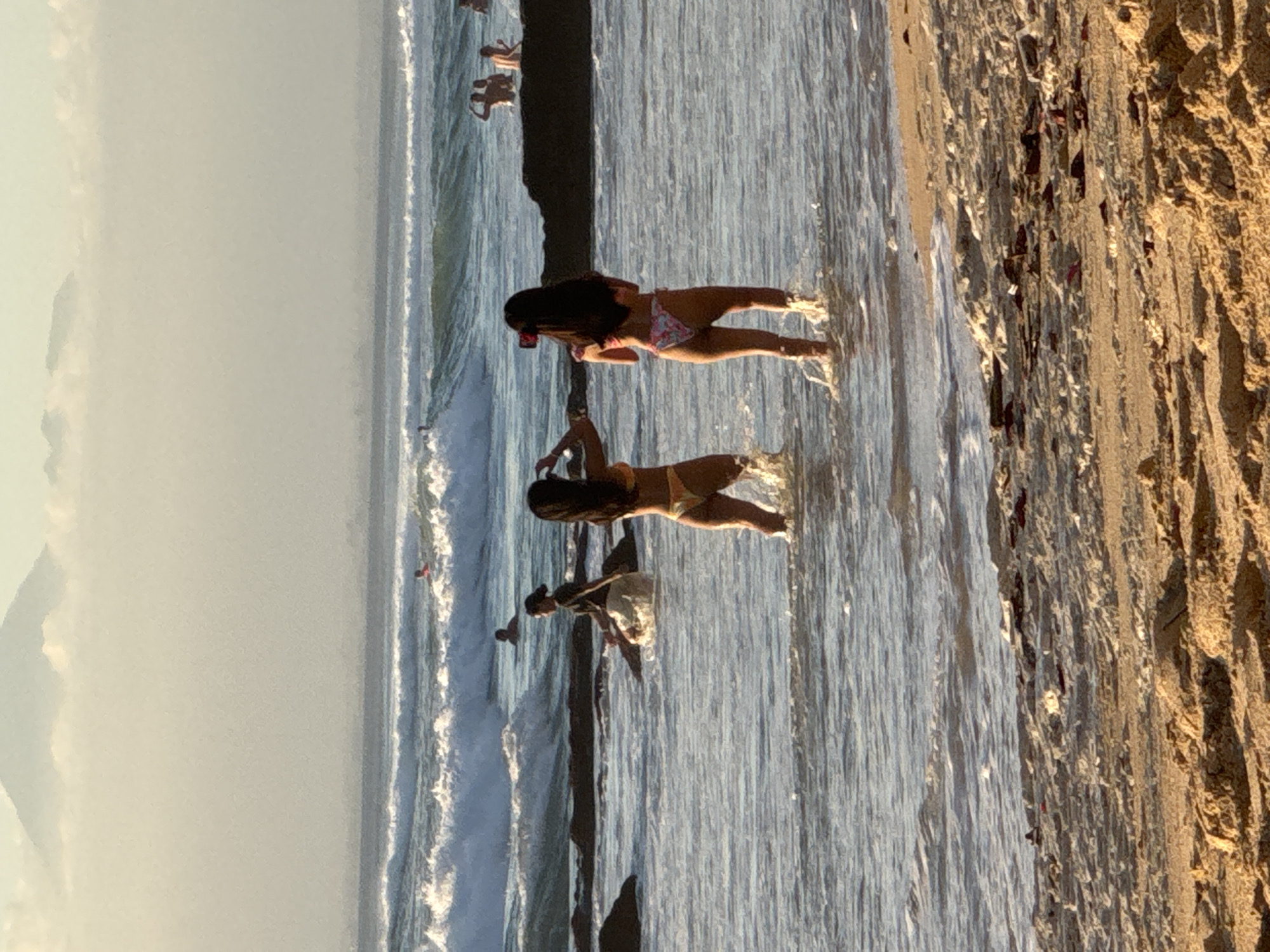 In Kuta Selatan, Indonesia, a beach scene unfolds with a hazy, mountainous backdrop. Two young women in bikinis stand in shallow water, one facing away from the camera, holding a phone to take a photo. The other woman, also facing away, poses. Waves crash on the shore, where a rocky outcrop is visible. In the distance, several people are swimming and surfing. The sand in the foreground is strewn with debris.