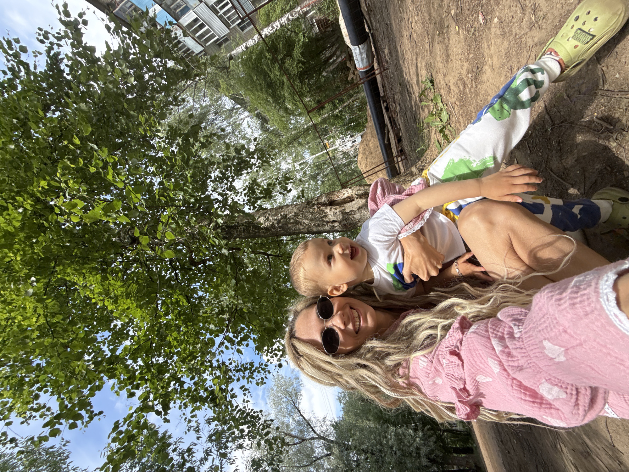 A vibrant, sunlit outdoor selfie captures a smiling woman and a young child, likely in Rusanovo, Russia. The woman, positioned closer to the camera, has long, wavy blonde hair and wears dark round sunglasses and a pink ruffled top adorned with white dots. She is smiling broadly. Nestled close to her, presumably being held, is a young child with blonde hair, also smiling and looking upwards. The child wears a white t-shirt with colorful abstract designs and white pants featuring green, blue, and yellow patterns. On the child's foot, a lime green Croc is visible with the text "GAME OVER" printed on it.
The setting appears to be an urban or semi-urban green space, possibly a park or courtyard. The background is dominated by lush green trees with dense foliage against a clear blue sky with scattered white clouds, indicating a bright, sunny day. In the upper right corner, parts of a building with multiple windows and some industrial-looking metal pipes or structures are visible. The ground beneath the subjects appears to be natural earth or a light-colored surface. The clear skies and strong shadows suggest it is midday or early afternoon. Both the woman and child exhibit happy, warm expressions, conveying a joyful interaction.