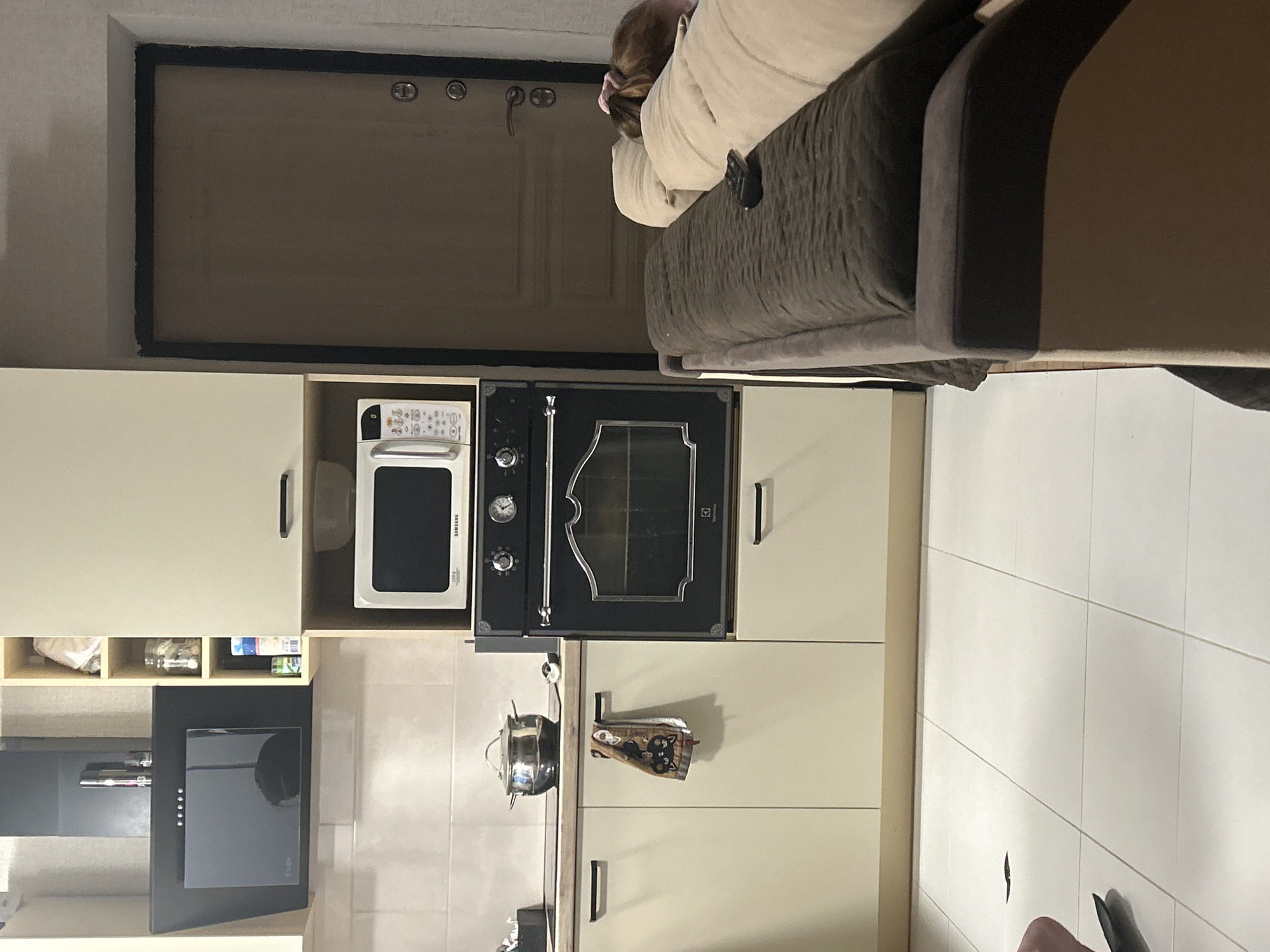 The image depicts an interior scene, likely a kitchen and living area, in an urban setting. In the kitchen area, a modern black range hood is mounted above a stovetop. To the right, a stack of kitchen cabinetry features a built-in oven and a microwave oven. A pot sits on the stovetop, and a towel hangs from a cabinet handle. The flooring in the kitchen is tiled in a light color.
Adjacent to the kitchen, a door with a textured surface and dark hardware is visible. To the right of the door, a sofa is positioned. The sofa is upholstered in a dark brown fabric, with lighter colored pillows on top. A remote control rests on one of the pillows. A person, partially visible, is lying on the sofa, with their head resting on a pillow.
The overall impression is of a domestic environment. The presence of kitchen appliances and a sofa suggests a living space where cooking and relaxation occur. The lighting appears to be artificial, casting shadows and contributing to a slightly dim atmosphere in some areas. The image does not provide any specific geographical markers that would definitively place it in Нижние Муллы, Russia; the description of the location is therefore speculative and based on the prompt's request.
