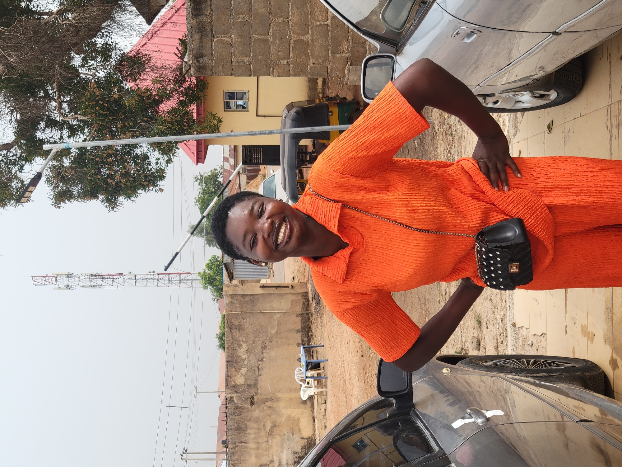 A woman with short black hair and an orange outfit is smiling at the camera. She is standing between two parked cars, a silver sedan and a darker-colored one. She has a small black purse hanging from her shoulder. Behind her is a building with a pink roof and another building with a yellow facade. A three-wheeled vehicle and a communication tower are visible further back. In the foreground, there is a paved area with a portion of a sidewalk visible. A section of the city of Gura Top, Nigeria, can be observed in the background.