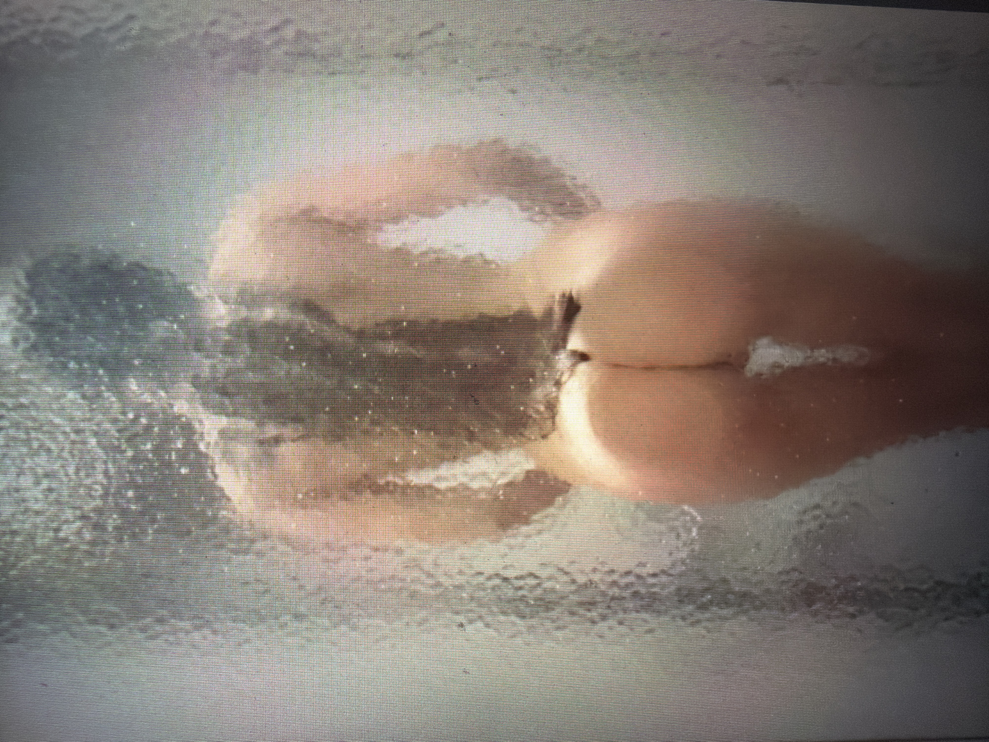 The image shows a close-up, horizontal view of a person from the back, partially submerged in water. The person's buttocks are prominently visible, with skin appearing smooth and peachy. The water is frothy and appears to be bubbly, suggesting it could be a bath or a shower. The lighting is soft and warm, creating a serene and intimate atmosphere. The background is blurred and out of focus, with hints of diffused light creating a hazy effect. The overall impression is one of relaxation and personal care, with the focus on the textures of water and skin. There is no visible text in the image.