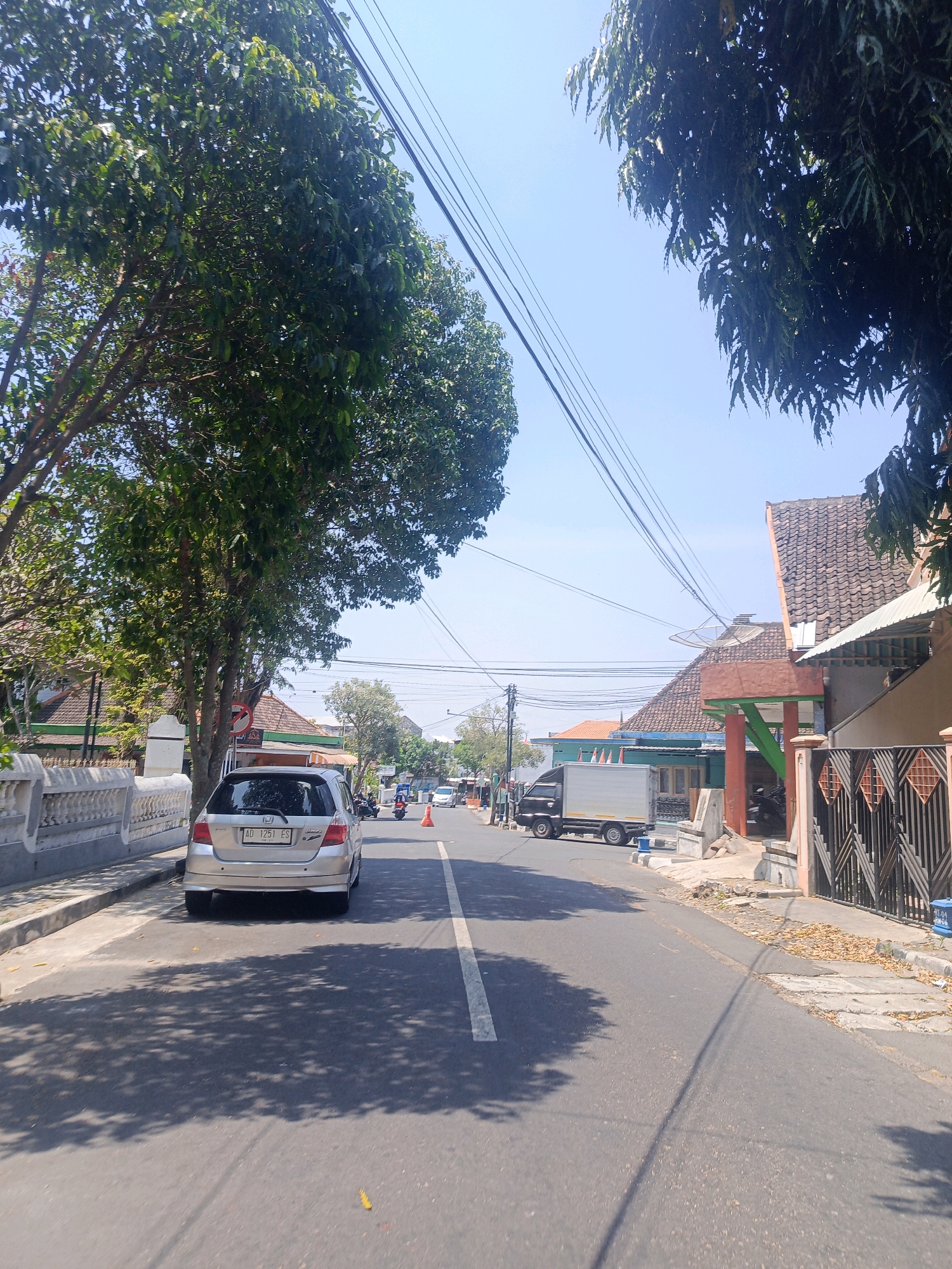 The image shows a street scene in Kebonagung, Indonesia. A silver Honda Jazz is parked on the left side of the road. A white truck with a silver cargo container is driving on the right side of the road. Several houses and trees line the street. The sky is clear and blue. The weather appears to be sunny and warm. There are power lines running overhead.