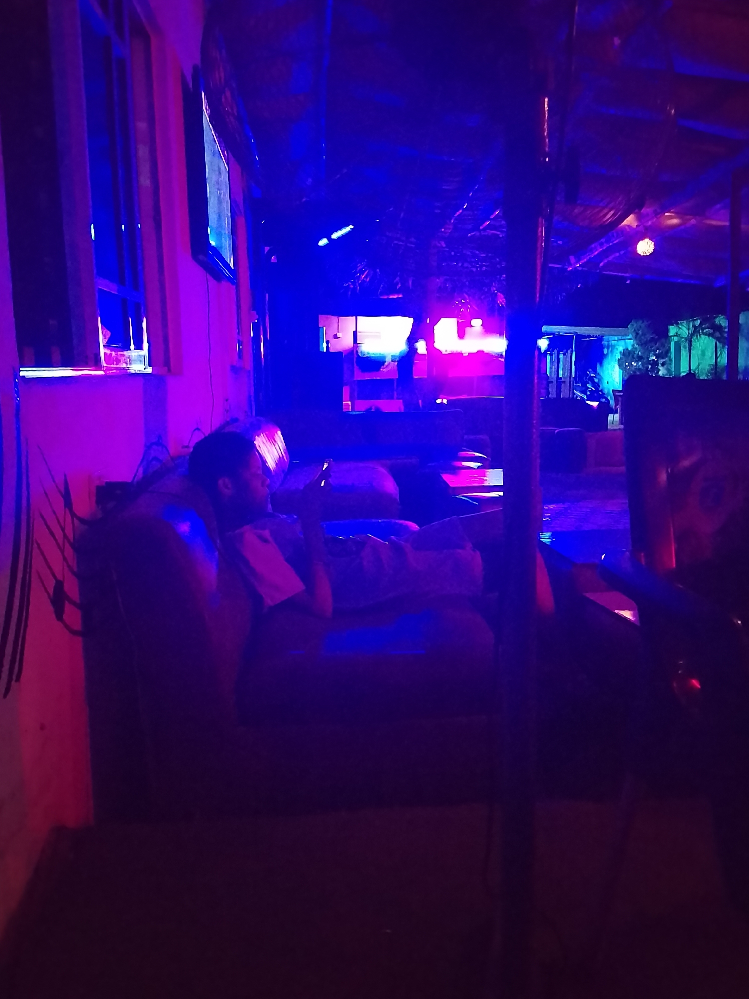 A person is sitting in an armchair indoors, looking at a handheld device. The lighting is predominantly blue and pink. The scene appears to be inside a bar or lounge. There is a window with blinds in the background, and a support pole runs vertically in the right foreground. Through the open area, there appear to be plants and outdoor lighting.