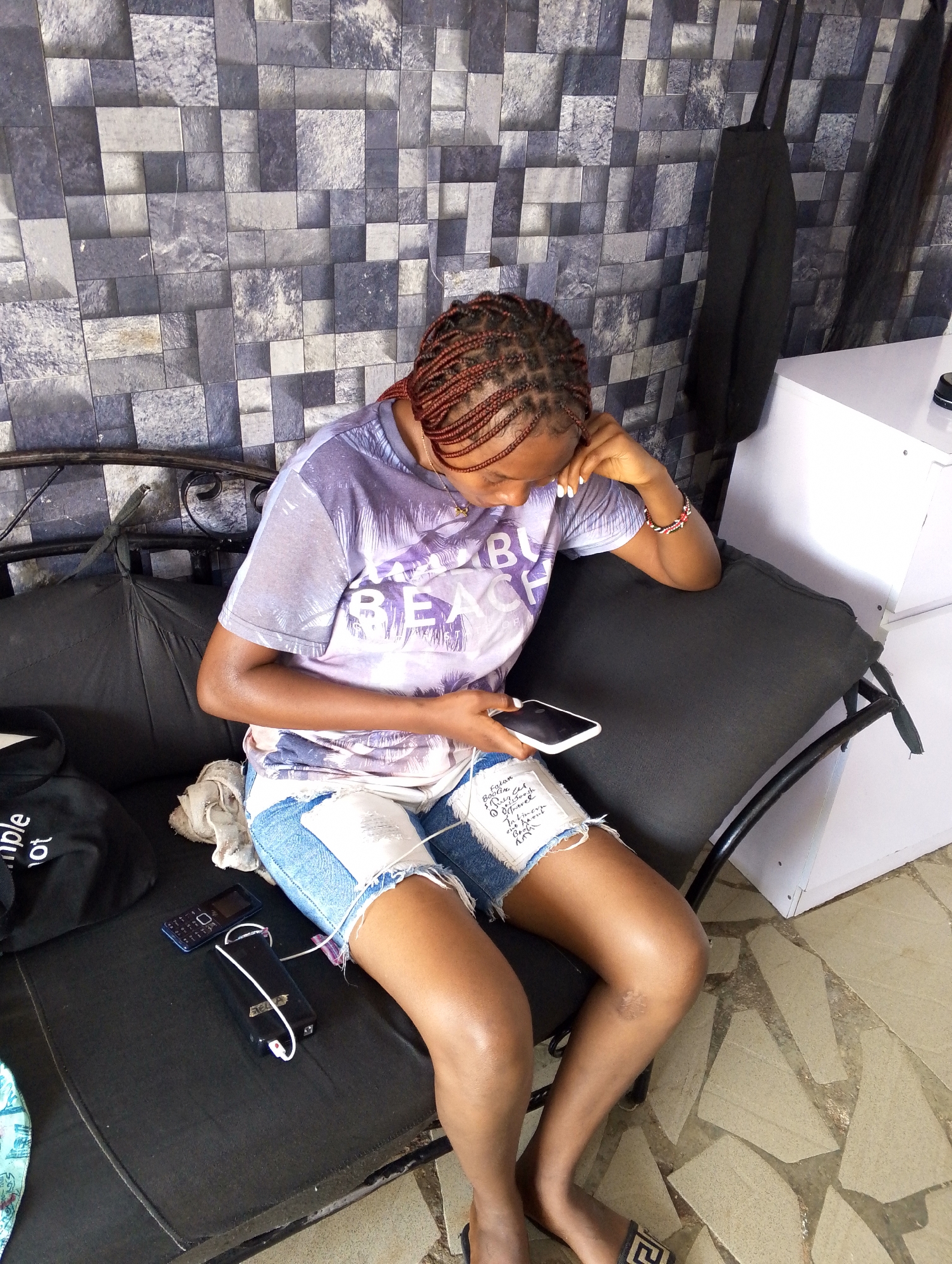 A young Black woman with braided auburn hair sits on a dark-colored outdoor sofa against a gray tiled wall. She is wearing a light purple t-shirt with a palm tree graphic and denim shorts with ripped detailing. She is looking down at her phone, which she holds in her right hand. Her left arm rests on her knee, and her left hand is near her face. A small black flip phone and a black power bank with a white charging cable are on the sofa next to her left leg. A black bag with white lettering is visible on the left side of the sofa. The floor is a light brown stone tile pattern.