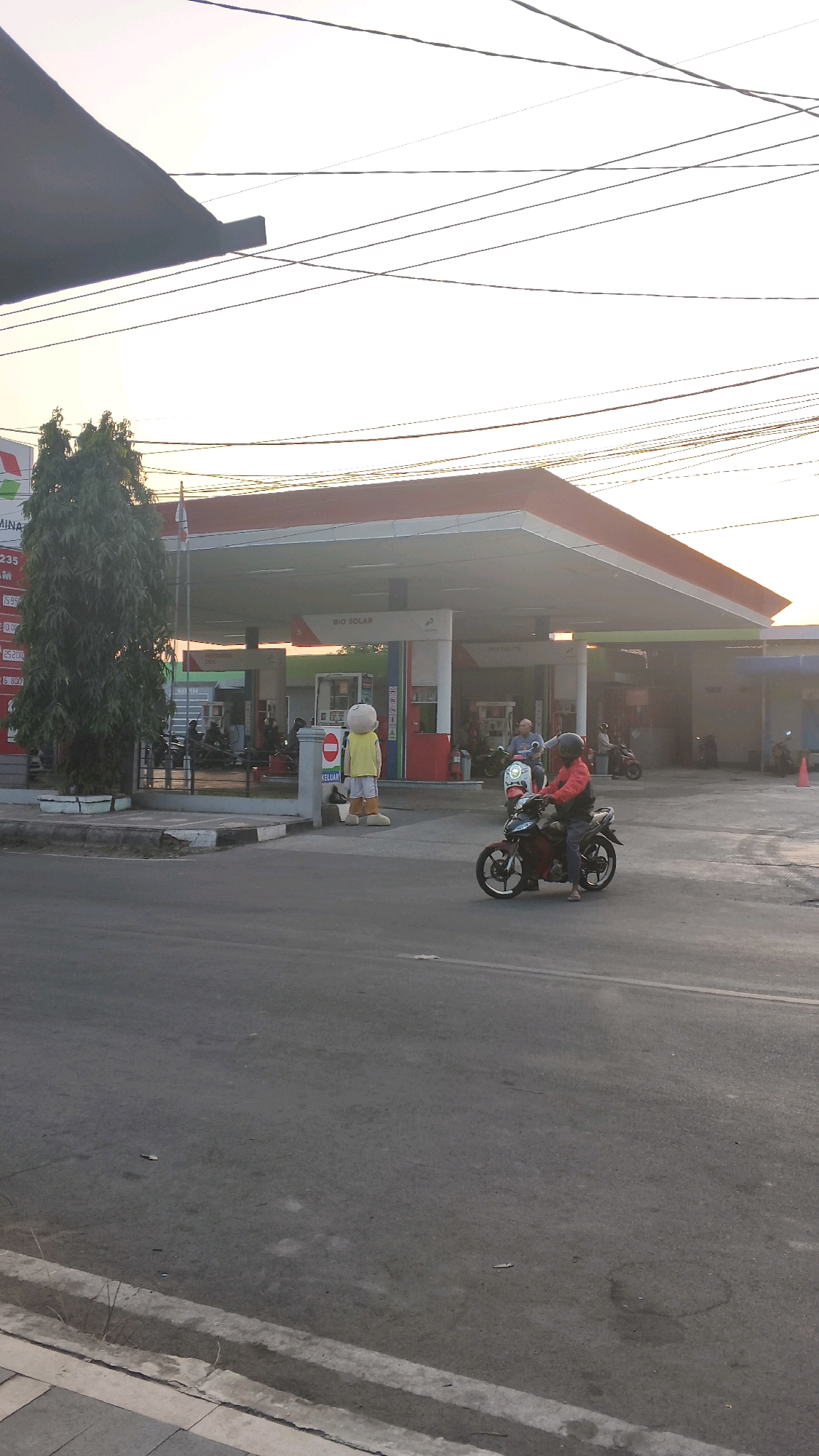 The image depicts a gas station in Margadadi, Indonesia, during what appears to be sunset. A Pertamina gas station with a red and white canopy is visible, featuring prominent signage indicating "BIO SOLAR" and "PERTALITE." Several fuel pumps are lined up under the canopy. In front of the station, a costumed character wearing a yellow shirt and shorts stands near a "KELUAR" (exit) sign.
On the asphalt road in front of the gas station, two individuals are riding motorcycles. One motorcycle, closer to the foreground, is ridden by a person wearing a red jacket and a helmet, while the other motorcycle, further back, is ridden by a person in a blue shirt and a helmet. Other motorcycles are parked near the gas station, and several people are visible near the pumps. A tall, mature tree with dense foliage stands to the left of the gas station, adjacent to a white planter. Power lines crisscross the sky above the scene. The overall lighting suggests the warm, soft glow of the setting sun.