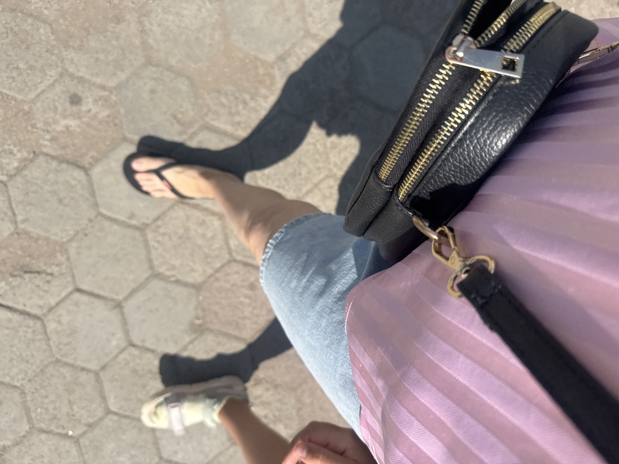The image shows a close-up, overhead view of a person's legs and a portion of their attire, taken on a paved surface composed of hexagonal concrete tiles. The person is wearing light-colored sneakers on their left foot and black flip-flops on their right foot. They are also wearing a light blue denim skirt or shorts. In the foreground, a black leather bag with gold zippers is visible, resting on a pleated pink fabric, possibly a skirt or dress. The lighting suggests it is daytime, with distinct shadows cast by the person's legs. The scene appears to be an outdoor urban environment, likely a sidewalk or pedestrian area. There is no visual information in the image to specifically identify the location as Kokshetau, Kazakhstan.