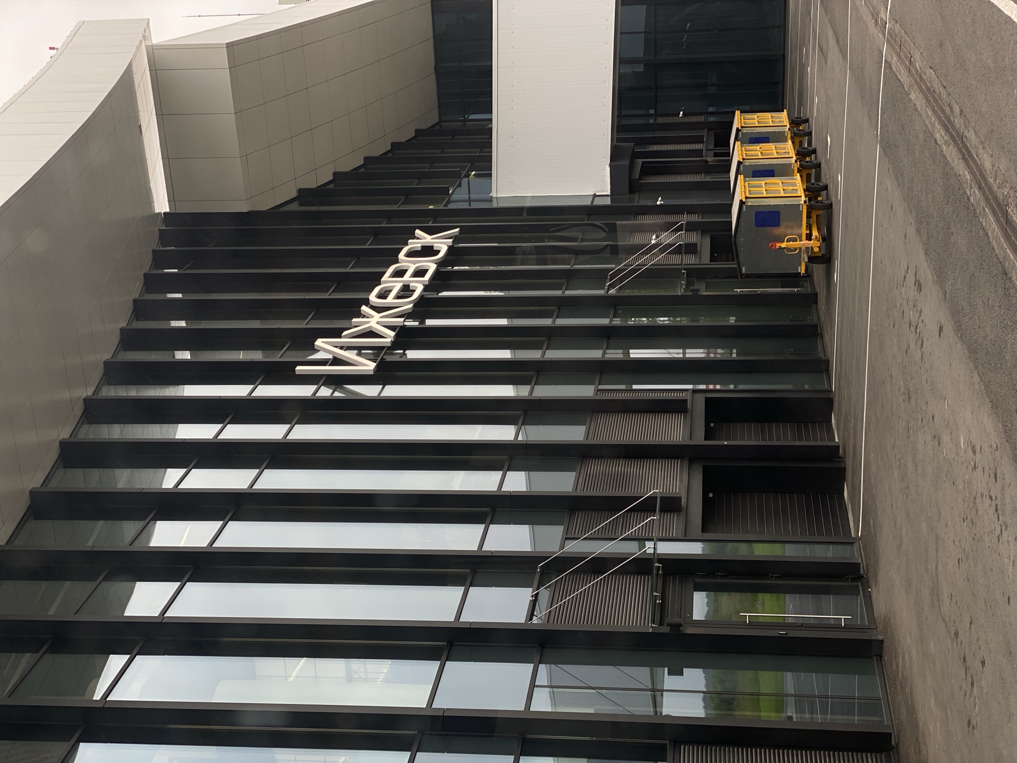 The image shows the exterior of a modern building with large glass windows and dark vertical panels. Large, illuminated white letters spelling out "ИЖЕВСК" are mounted on the facade. In front of the building, a paved area shows white parking lines and a drain. Lined up in a row are three identical small, yellow-and-gray wheeled carts. The building appears to be an airport terminal or similar public facility, given the architecture and the presence of baggage carts. The surrounding environment is not clearly visible, but glimpses of greenery suggest an outdoor setting. The location is identified as Novoye Martyanovo, Russia.