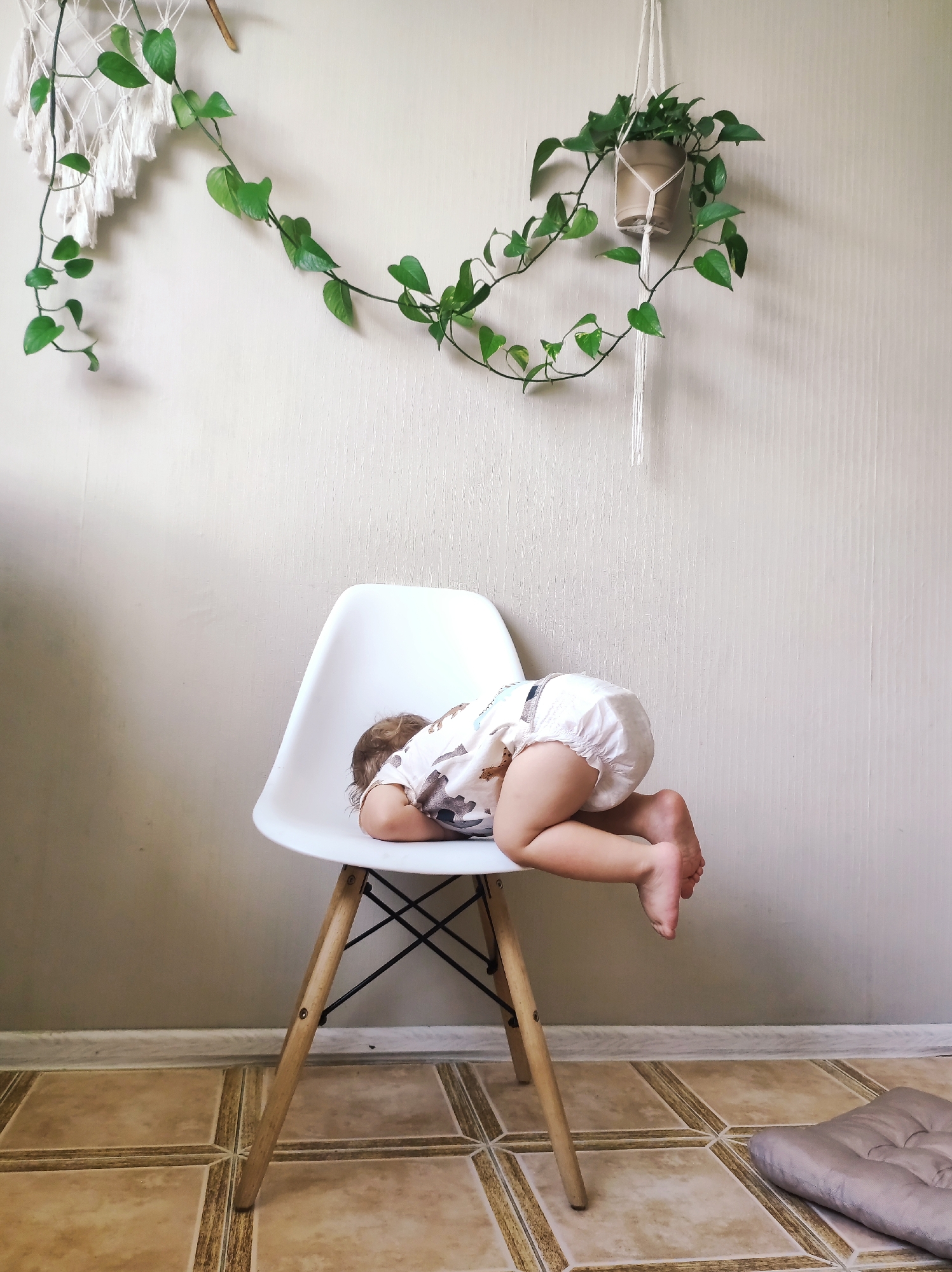A baby is sleeping on a white chair in a room. The baby is wearing a diaper and a light-colored shirt with a pattern. The baby's head is resting on their arms, and their legs are dangling over the side of the chair. The chair has wooden legs and a black cross brace. The floor is tiled with light brown tiles. In the background, there is a light-colored wall with a textured pattern. Two hanging plants are visible on the wall, one with tassels and the other in a pot. The overall scene appears to be indoors, possibly a home. The lighting suggests it could be daytime, with natural light coming from an unseen source. The mood is peaceful and serene due to the sleeping baby. There is no visible text in the image.