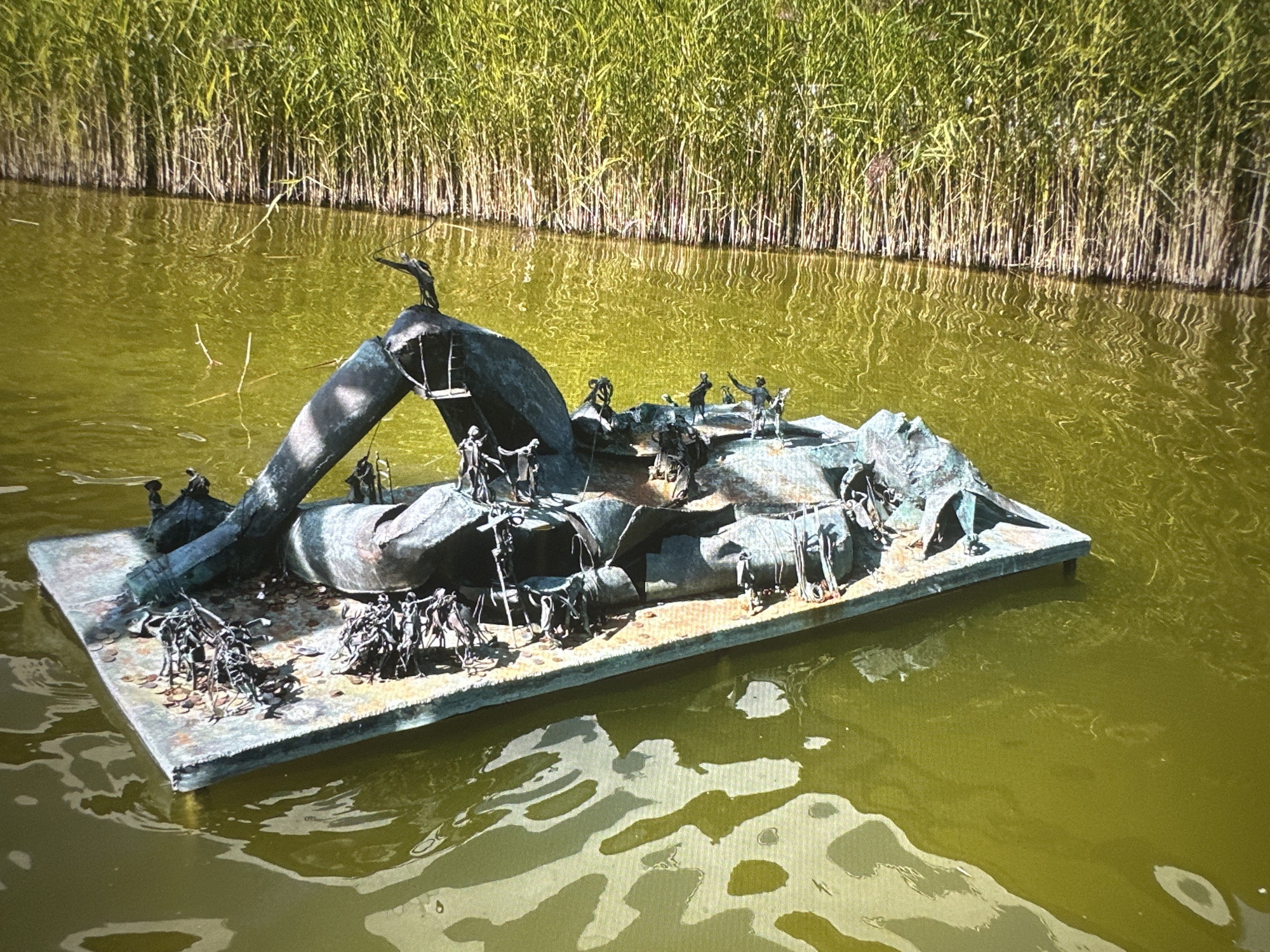 A bronze sculpture is floating on a murky green body of water. The sculpture depicts a collection of small human figures interacting with a larger, abstract form that resembles a fragmented body or structure. Some figures appear to be climbing, reaching, or engaged in other activities on and around this form. The sculpture rests on a rectangular base that is partially submerged in the water. In the background, a dense wall of tall green reeds lines the edge of the water. The water's surface is disturbed by ripples, reflecting the reeds and the sky. The lighting suggests it is daytime, with sunlight illuminating the scene. The location is described as Unknown, Unknown.