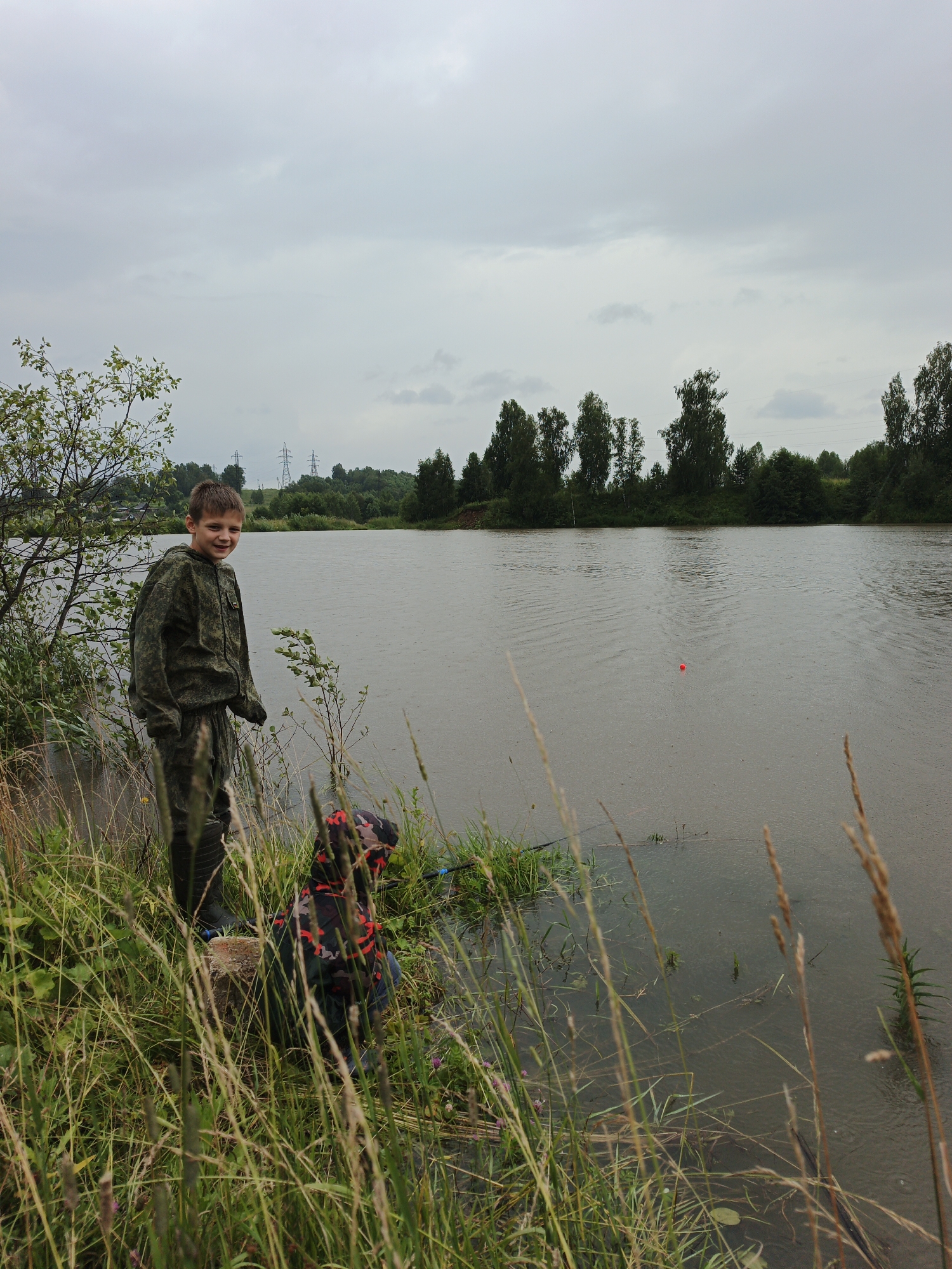 In Urtiushka, Russia, a boy in camouflage fishing attire stands on the grassy bank of a body of water. The boy smiles at the camera, his fishing gear visible. Nearby, a younger child, also in camouflage, is crouched down, facing away from the viewer and towards the water. The scene is set against a backdrop of trees and a cloudy sky, with power lines visible in the distance. The water surface shows ripples, suggesting it is either raining or windy.