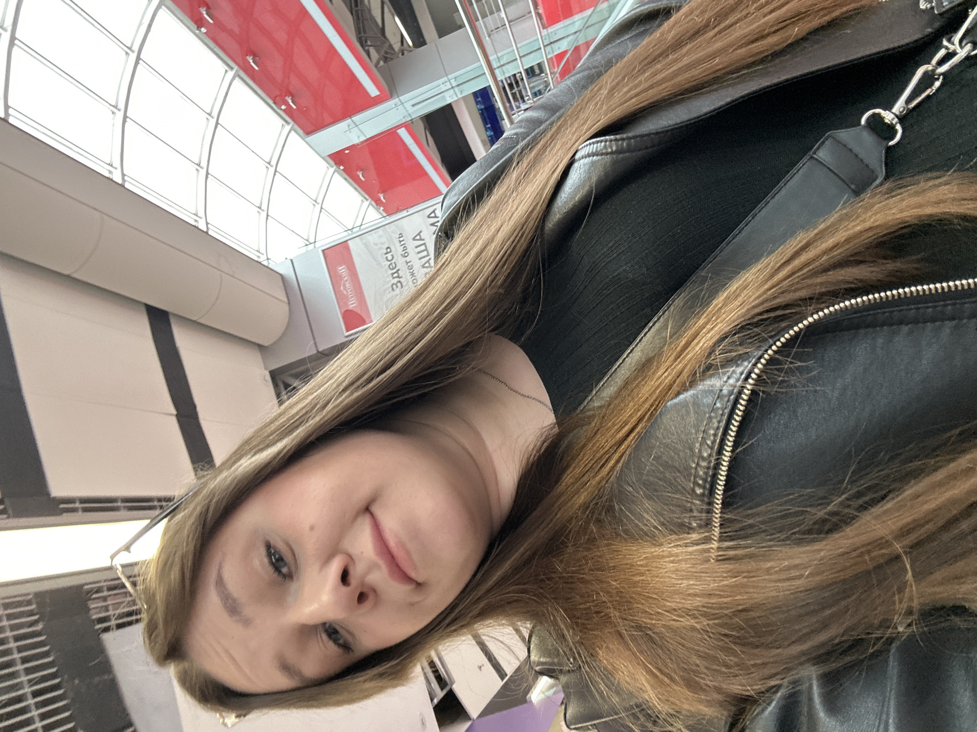 A woman with long, light brown hair is the primary subject. She is wearing a black top and a black leather jacket with a silver zipper detail. A black strap with a silver clip is visible on the right side of the frame. She appears to be indoors in a location in Izhevsk, Russia, as indicated by a sign in Russian visible in the background. The background also includes parts of the building's architecture, including a white ceiling with a curved, windowed section and red and white rectangular panels above it.