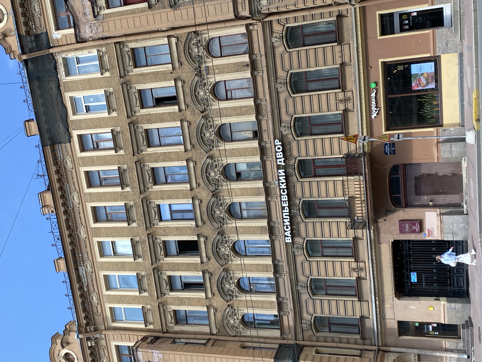 This media file captures a street view of a building in Saint Petersburg, Russia. The building's facade is constructed of brick and decorated with ornate architectural details, including moldings and sculptures around the windows. The windows themselves are large and rectangular, some with shutters, and others are arched. The overall style suggests historical architecture.
On the right side of the building, there are businesses with storefronts. One storefront has a sign with white lettering that reads "КОРЕЯНА" and displays a digital screen showing food. Another sign above it, in a dark rectangular frame, displays smaller icons. Below this, there is an entrance with a black metal gate. A person, dressed in light-colored clothing, is walking through the gate, with their arm outstretched.
Visible text includes "КОРЕЯНА", "РИМ", and what appears to be street signage with numbers and Cyrillic text. The scene is illuminated by natural light, suggesting daytime, and the sky is visible and blue, indicating fair weather. The presence of businesses and street signs confirms an urban setting. The architectural style and visible text provide strong cues to the location being Saint Petersburg, Russia.