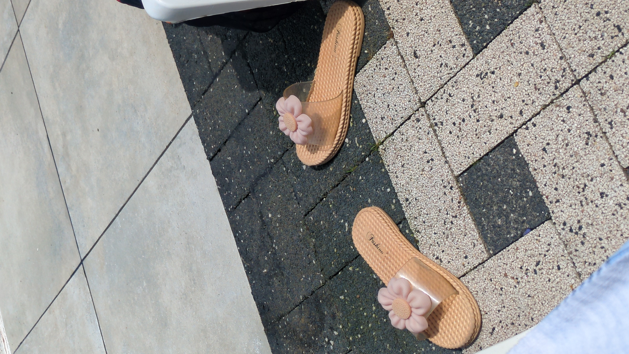 Two light pink slides adorned with flower embellishments rest on a textured, interlocking pavement. The slides are positioned parallel to each other, with the left slide slightly closer to the viewer. The pavement consists of a mix of dark grey and light speckled concrete blocks. In the background, a strip of plain grey concrete is visible, separated from the darker pavement by a thin line. A white object, possibly a piece of furniture, is partially visible on the right edge of the frame. The scene appears to be an outdoor setting, likely a patio or poolside area, under bright sunlight, casting subtle shadows.