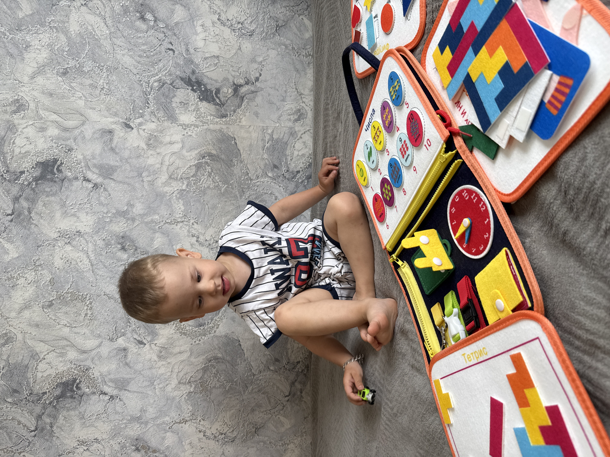 A child wearing a striped shirt sits on the floor next to an open, interactive felt book. The book features educational activities, including number and shape matching, a clock face, and zipper closures. The floor is a two-toned gray, and the setting appears to be inside a home. Rusanovo, Russia is not evident in the image.