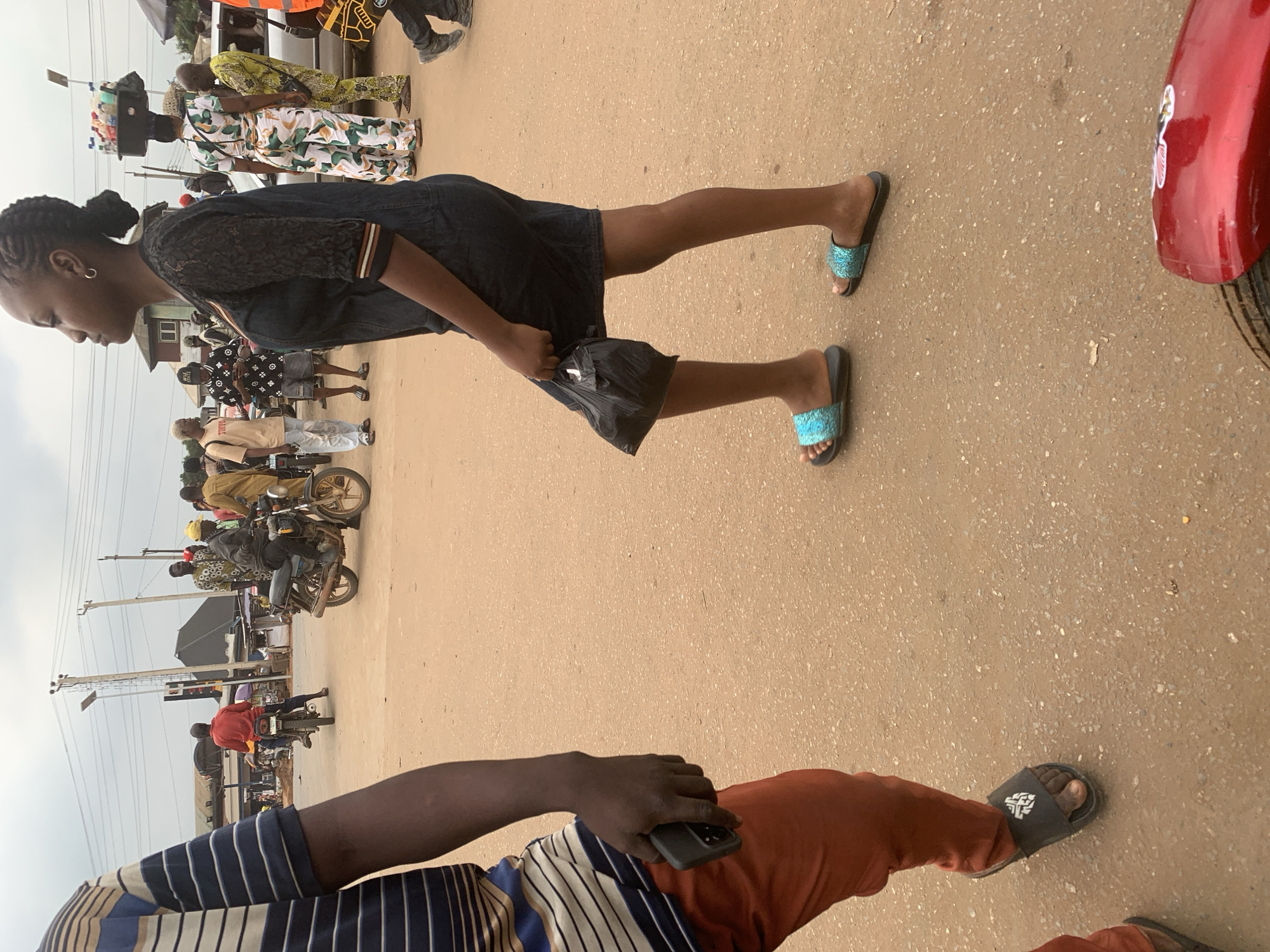 In Shagamu, Nigeria, a street scene unfolds with several individuals and vehicles. In the foreground, a person wearing an orange trousers and a blue and white striped t-shirt is partially visible, holding a smartphone. To their right, a woman in a dark dress walks, carrying a black plastic bag. Further in the background, a woman wearing a patterned outfit carries a stack of items on her head. Various motorcycles are present, with some people riding them and others standing nearby. A red motorcycle fender is visible in the bottom right of the image. The environment appears to be a dusty, unpaved street with buildings and utility poles in the background. The sky is overcast.