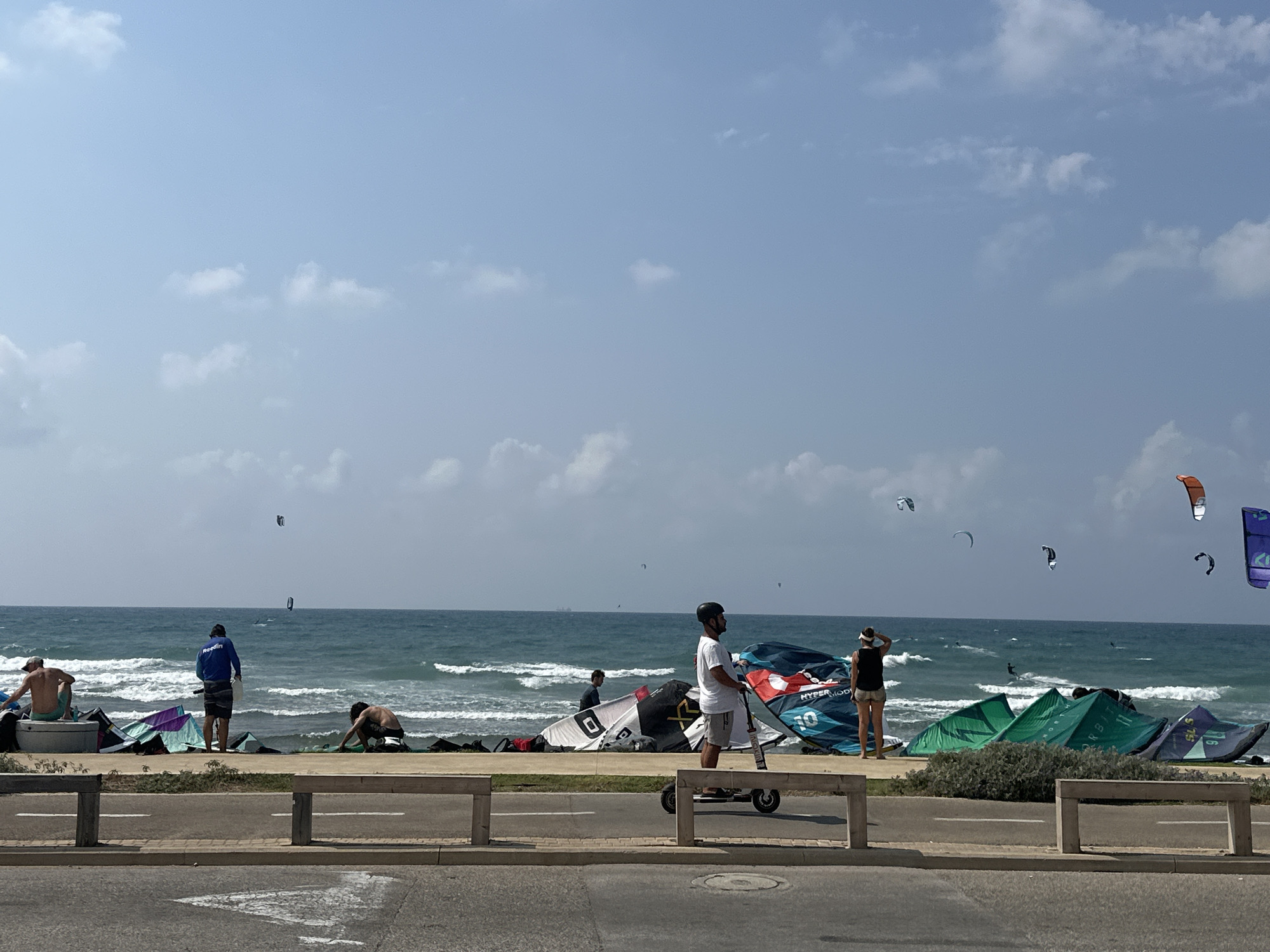 This image captures a breezy day at a beach in Haifa, Israel, likely on the Mediterranean coast. The scene is dominated by the sea and sky, with a clear blue sky dotted with scattered white clouds. The water is a deep blue, with white-capped waves indicating a moderate wind. A number of kite surfers are visible in the water, some actively engaged in their sport with colorful kites in the air, while others appear to be preparing or resting.
On the beach, several large kites are laid out on the sand, suggesting a kiteboarding or kitesurfing location. There are a few people on the beach, some of whom are involved with the kites, while others are observing the water. One person is standing on an electric scooter, facing the sea. The beach is bordered by a paved walkway and a barrier, suggesting it is a public area. The overall impression is one of a popular spot for water sports enthusiasts on a pleasant day.