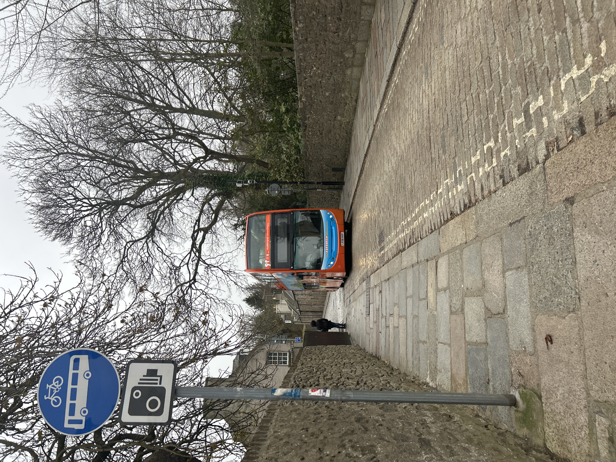 A slightly tilted, street-level shot captures a narrow, cobblestone street in Aberdeen City, United Kingdom. An orange and blue double-decker bus advances down the street toward the camera. A lone figure is walking in the same direction as the bus ahead of the bus. The street is flanked by stone walls on both sides. In the lower left corner, a sign attached to a metal pole shows a blue circular sign depicting a bus with a bicycle on top and a black and white rectangular sign featuring a camera. Bare trees frame the scene against a gray sky.