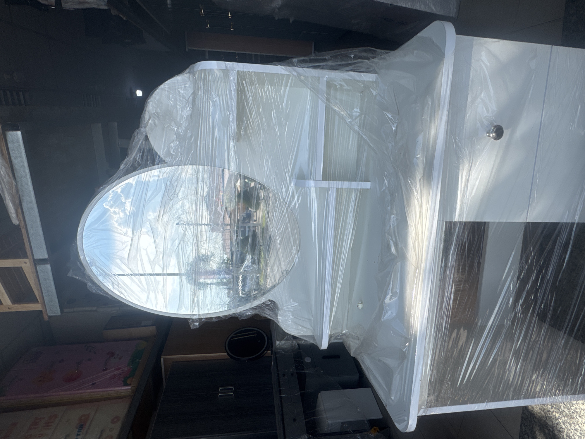 A white vanity with a large oval mirror is displayed in what appears to be a furniture store. The vanity and mirror are wrapped in clear plastic. The vanity has a smooth, white top with rounded edges and two drawers below. A single silver knob is visible on the top drawer. The mirror is framed by the same white material as the vanity and is attached to the back, curving slightly upwards. The reflection in the mirror shows a bright, cloudy sky and some buildings. To the left of the vanity, a dark wood cabinet with two doors is visible. Further to the left, stacked items wrapped in pink and clear plastic are on shelves. The overall environment suggests a retail setting, possibly a showroom or warehouse. There are no people visible in the image. The location is specified as Bac Nha Trang Ward, Vietnam, and the scene appears to be an interior of a commercial establishment.