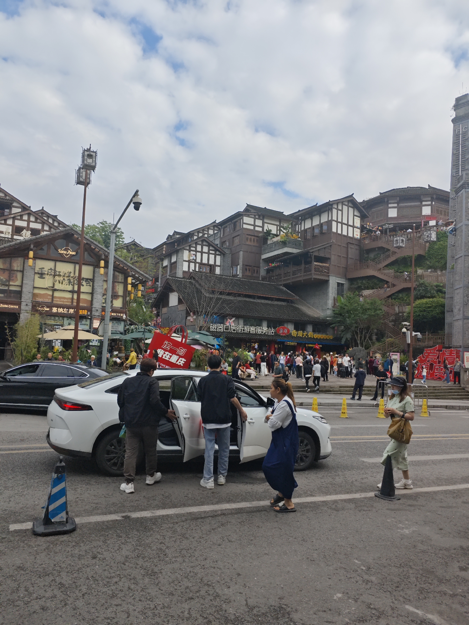 The image depicts a street scene in Shapingba District, China. In the foreground, a white car has its doors open, with two men standing next to it and a woman standing near the front of the vehicle. A woman in a light green dress and a face mask stands further to the right. Blue and white and plain black traffic cones are placed along the street.
Behind the car, a crowd of people is gathered. Buildings with traditional Chinese architectural elements, including wooden facades and tiled roofs, form the backdrop. A sign reading "Sana Zoan" and "ZOAN COFFEE" is visible on one building. A large red sign with Chinese characters is also present in the background.
Overhead, the sky is partly cloudy with patches of blue. A tall tower or structure is visible on the right side of the image. A black car is parked to the left.