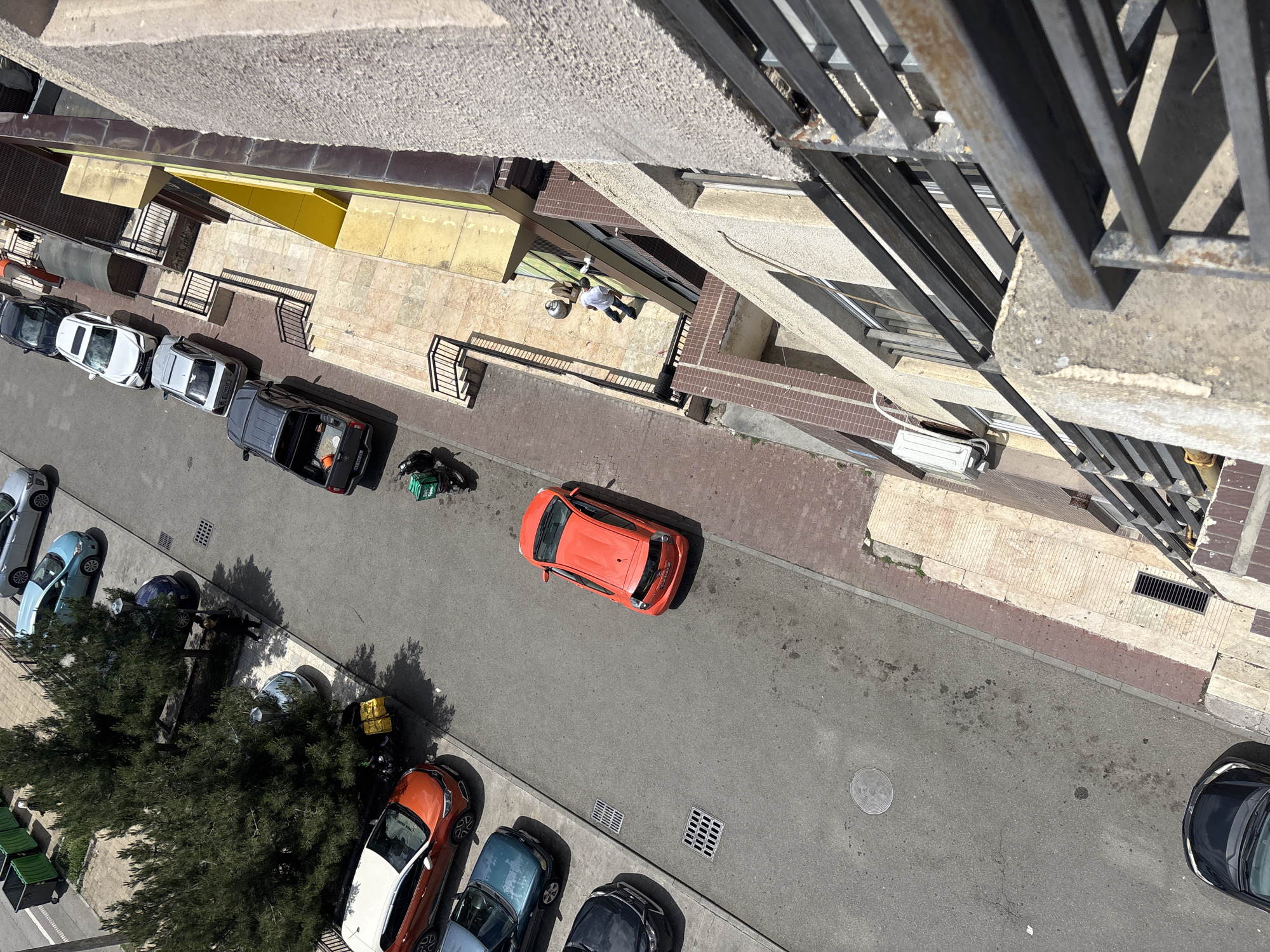 An aerial view, likely from a tall building, depicts a street scene in Tbilisi, Georgia. The street contains parked vehicles of various colors: white, silver, blue, black, and orange. An orange car is parked in the middle of the street. A black SUV has an open hatch. A person is laying on a patio. A person is standing next to a green delivery bag. Trees partially obscure the lower left corner of the frame, near more parked cars. Balconies are visible on adjacent buildings. A person is laying on a patio. A person is standing next to a green delivery bag.