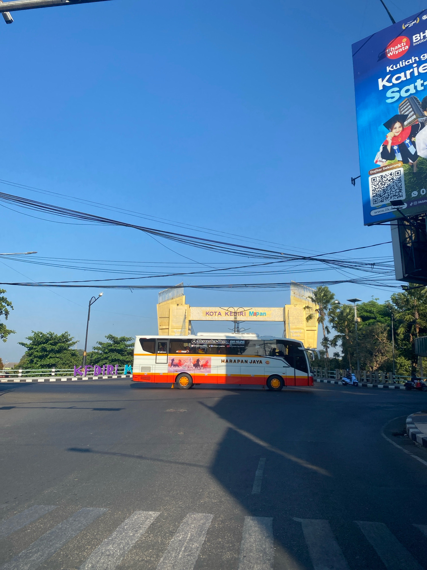The image captures a street scene in the city of Kediri, Indonesia. In the foreground, a bus with "HARAPAN JAYA" written on its side is driving on a road. The bus has orange and white coloring with graphics of horses on its side. Behind the bus, a large yellow archway is visible with "KOTA KEDIRI Mapan" written on it, indicating the location. Several utility wires are strung across the sky. To the right, a large billboard is mounted on a pole, featuring an advertisement with text in Indonesian. The sky is clear and blue. In the background, trees and palm trees are present. The road has white markings for a pedestrian crossing.