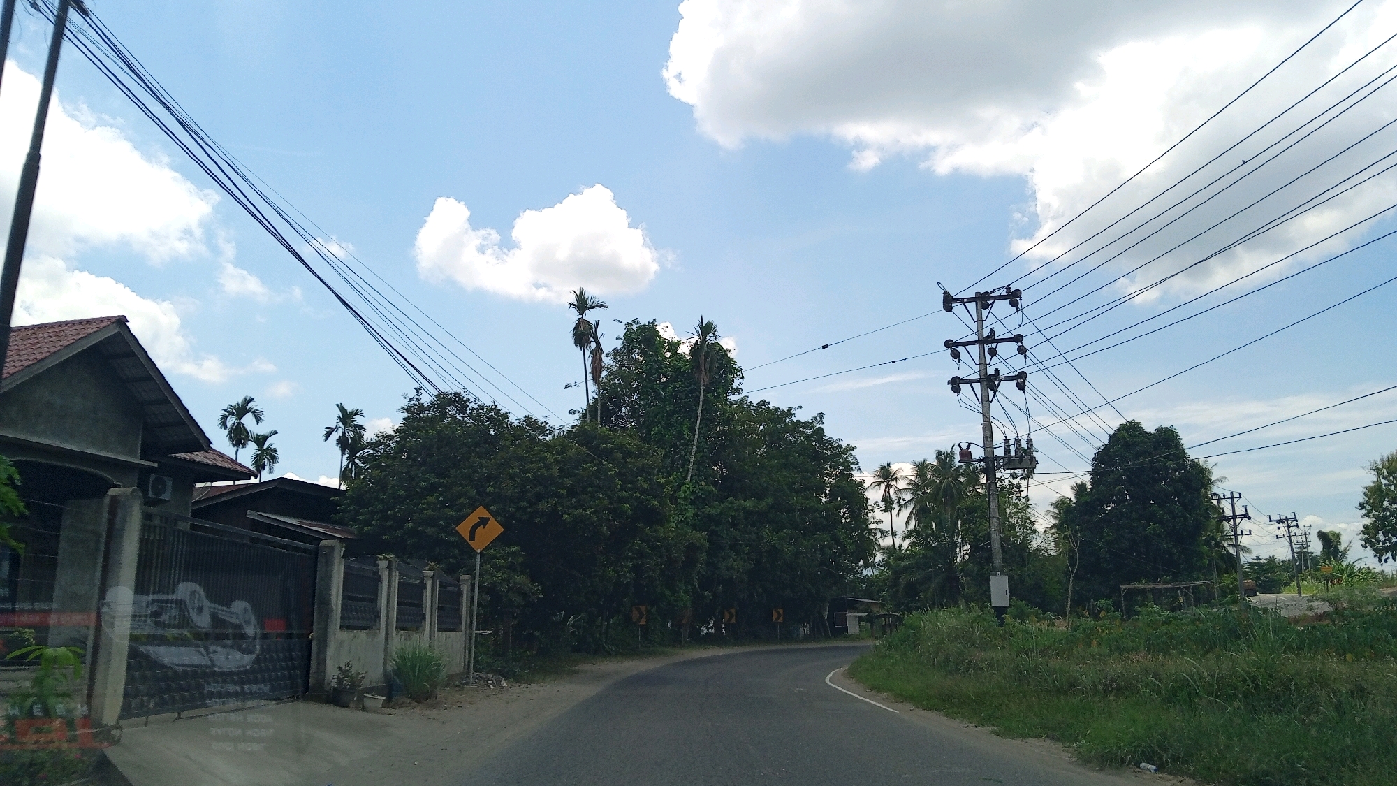The image captures a view of a curved asphalt road in Beunyot, Indonesia, under a bright blue sky with scattered white clouds. A residential building with a red tiled roof and grey walls is visible on the left, featuring a tall fence with a metal gate. The gate has a large, white graphic of a car or vehicle. Several utility poles with numerous electrical wires stretch across the sky. Dense green trees and palm trees line the right side of the road, with a few smaller buildings and more utility poles visible in the distance. A yellow diamond-shaped road sign with a black arrow indicating a curve is posted on the left side of the road. The overall scene suggests a rural or suburban setting.